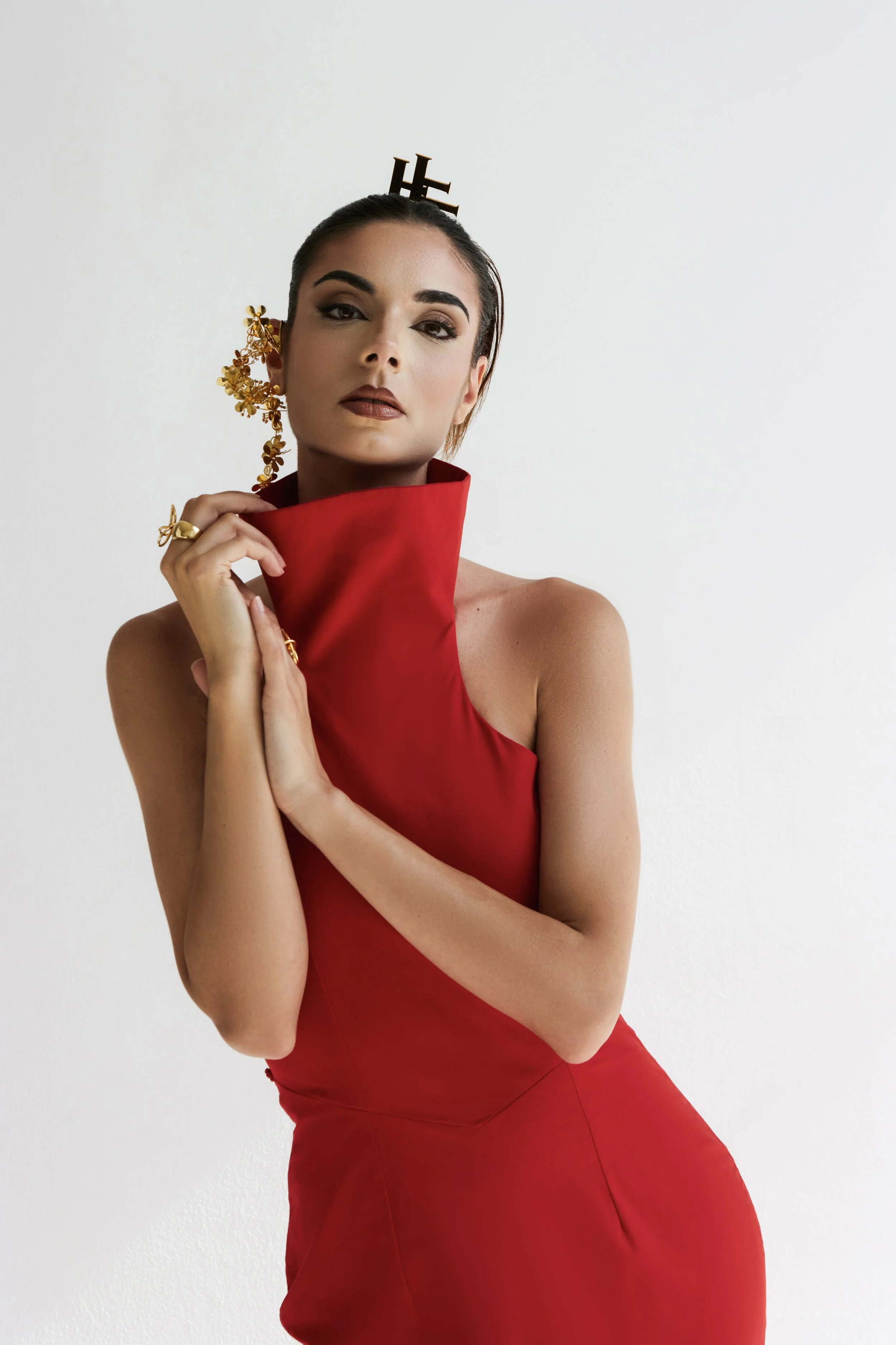 A woman in a red dress with gold jewelry and a stylized gold earring, holding the collar of her dress with one hand, stands against a plain white background.