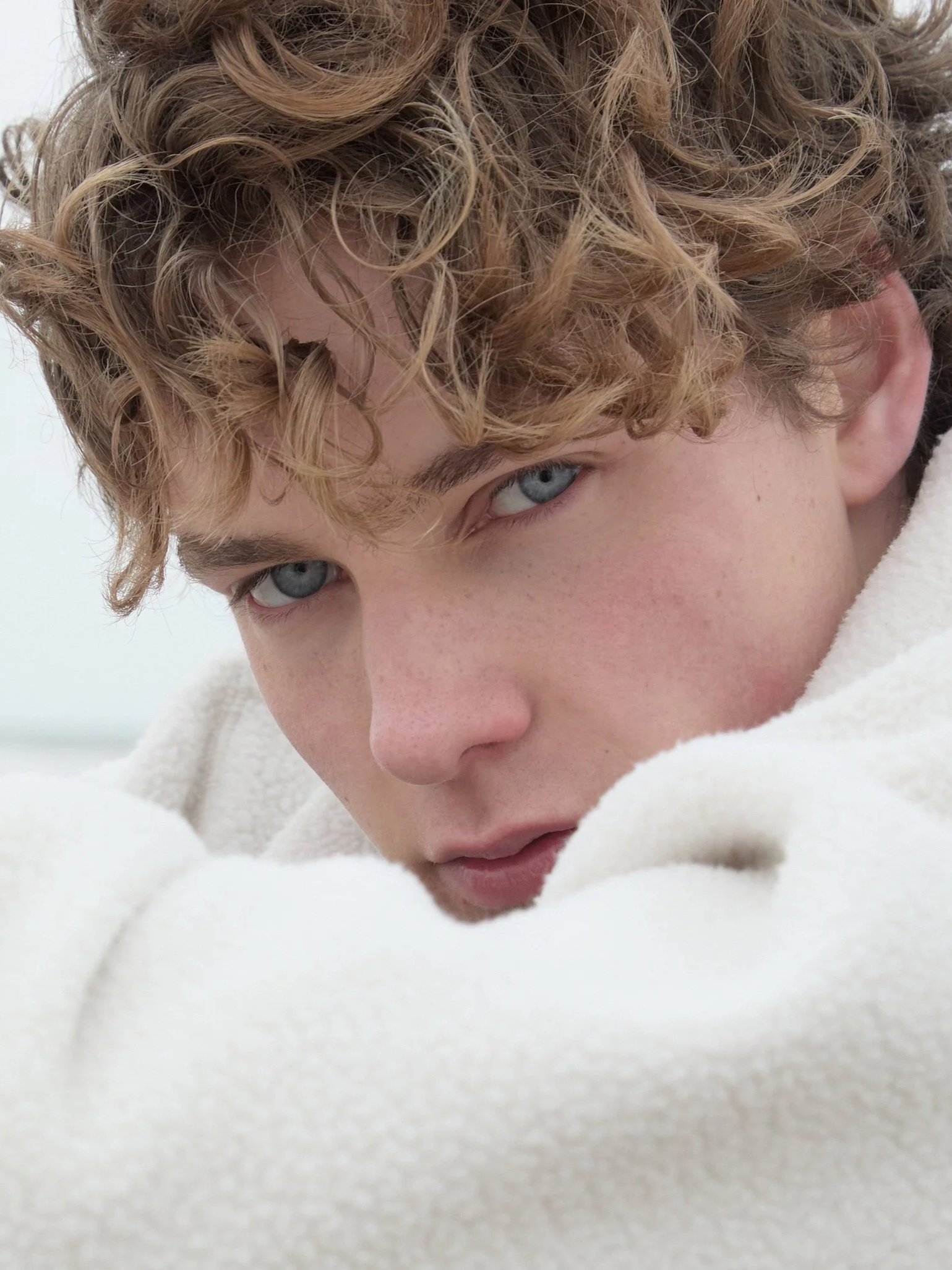 Close-up of a young person with curly blond hair and blue eyes, wearing a cream-colored fleece jacket, looking into the camera.