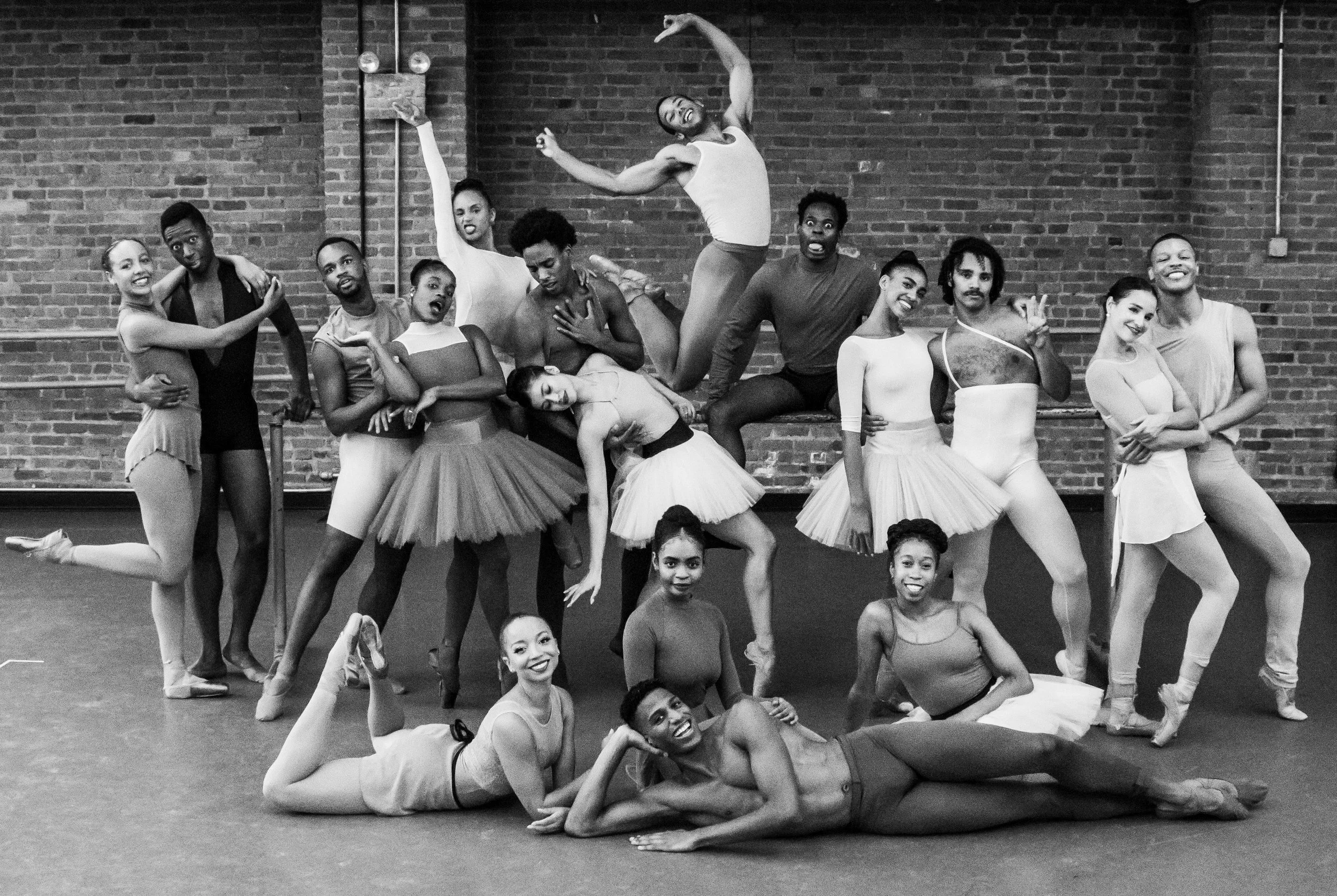 Black and white photograph of a diverse group of ballet dancers and choreographers in a dance studio posing for a group photo, with a brick wall in the background and some of the dancers in ballet costumes and tights.