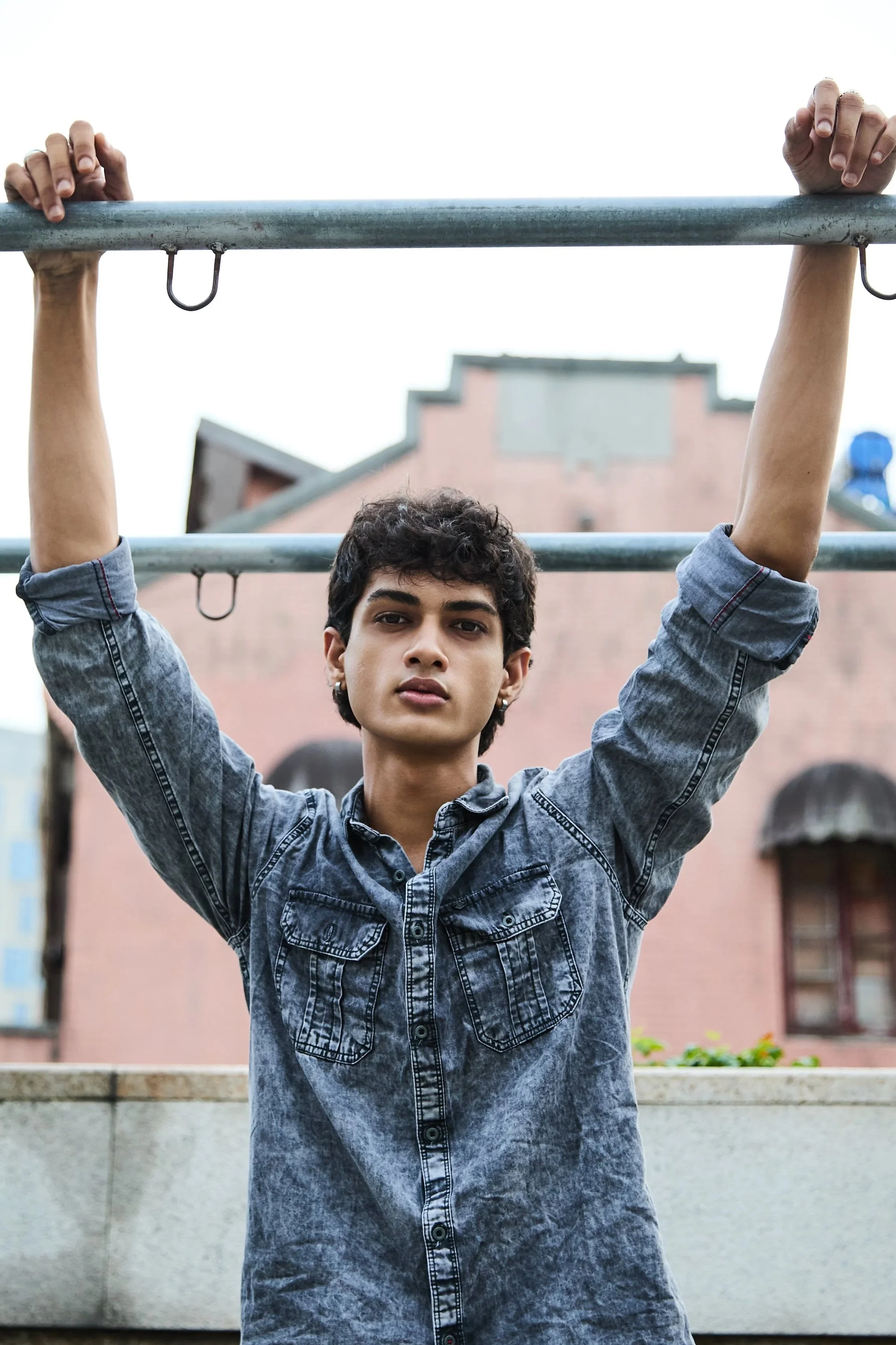 A young man with dark curly hair holding onto a horizontal bar with both hands, standing outdoors in an urban setting with buildings in the background.