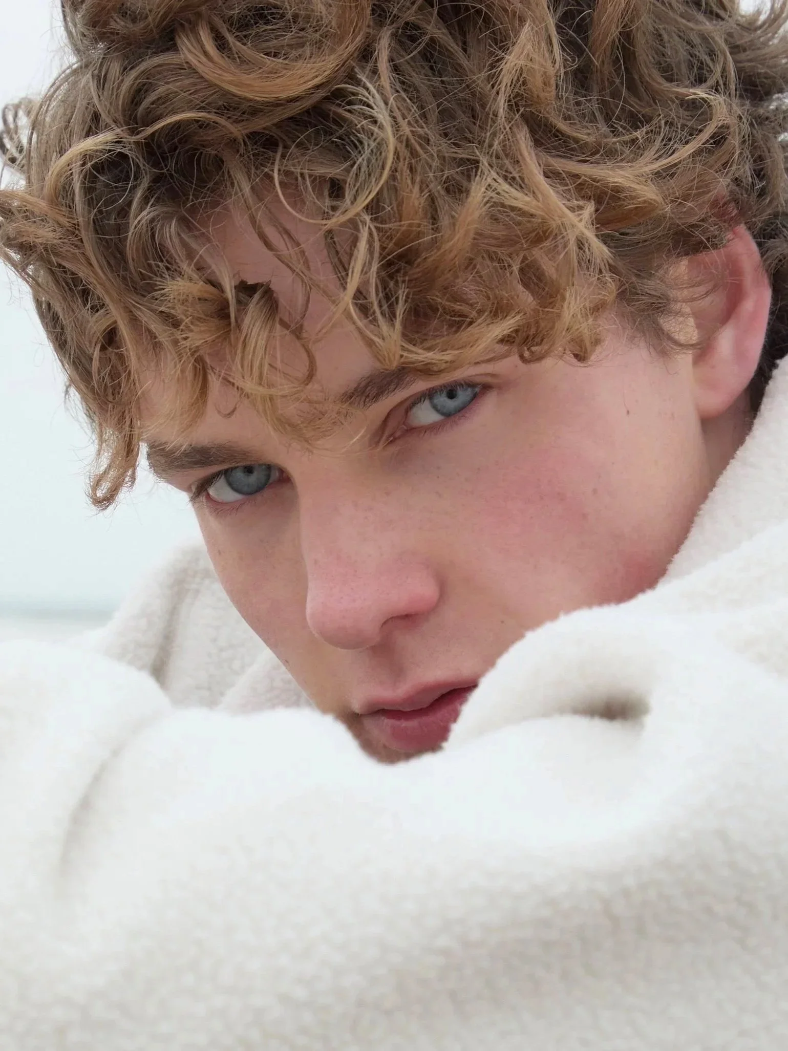 Close-up of a young person with curly blond hair and blue eyes, wearing a cream-colored fleece jacket, looking into the camera.