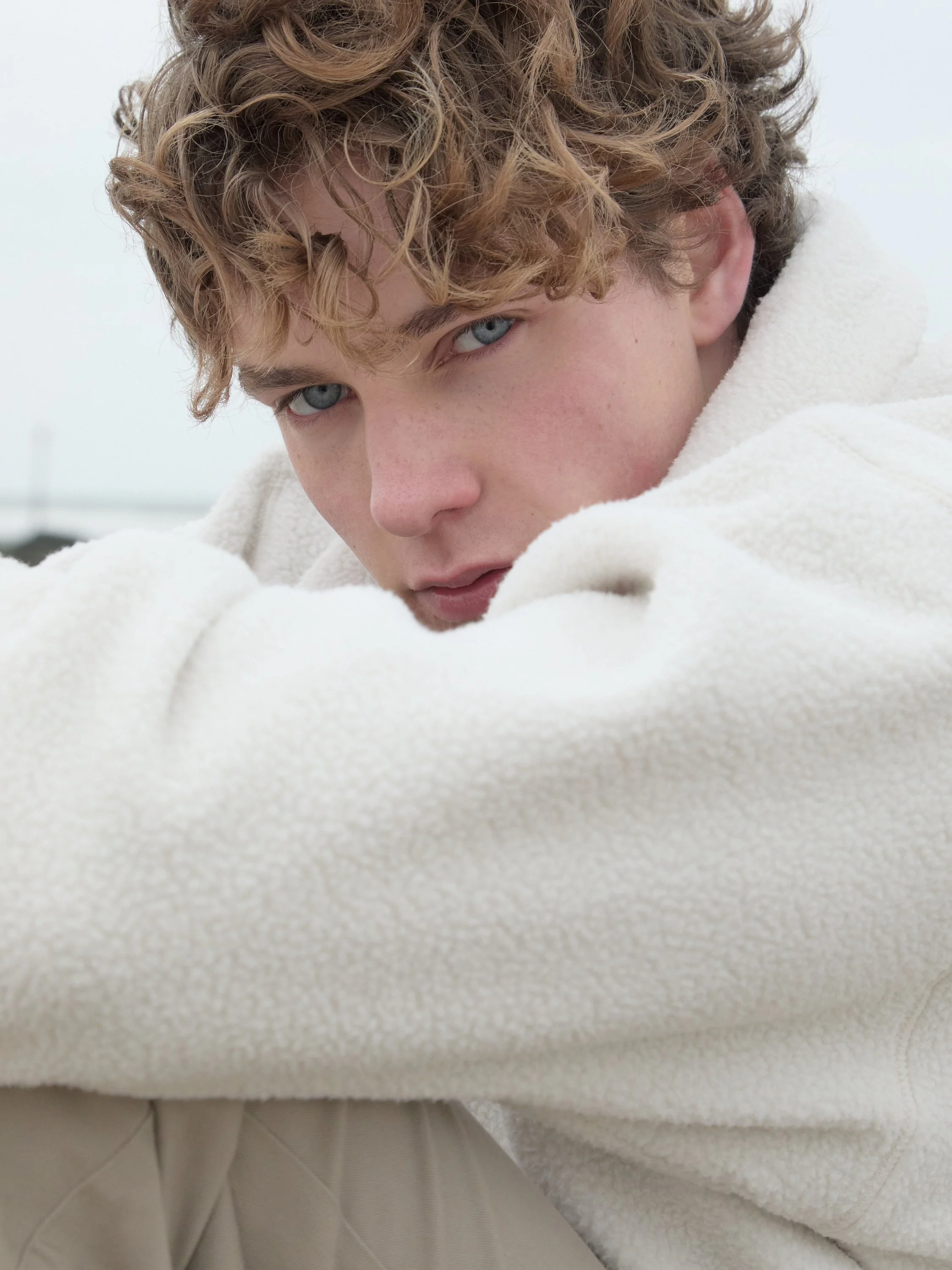 Close-up of a young person with curly blond hair and blue eyes, wearing a cream-colored fleece jacket, looking into the camera.