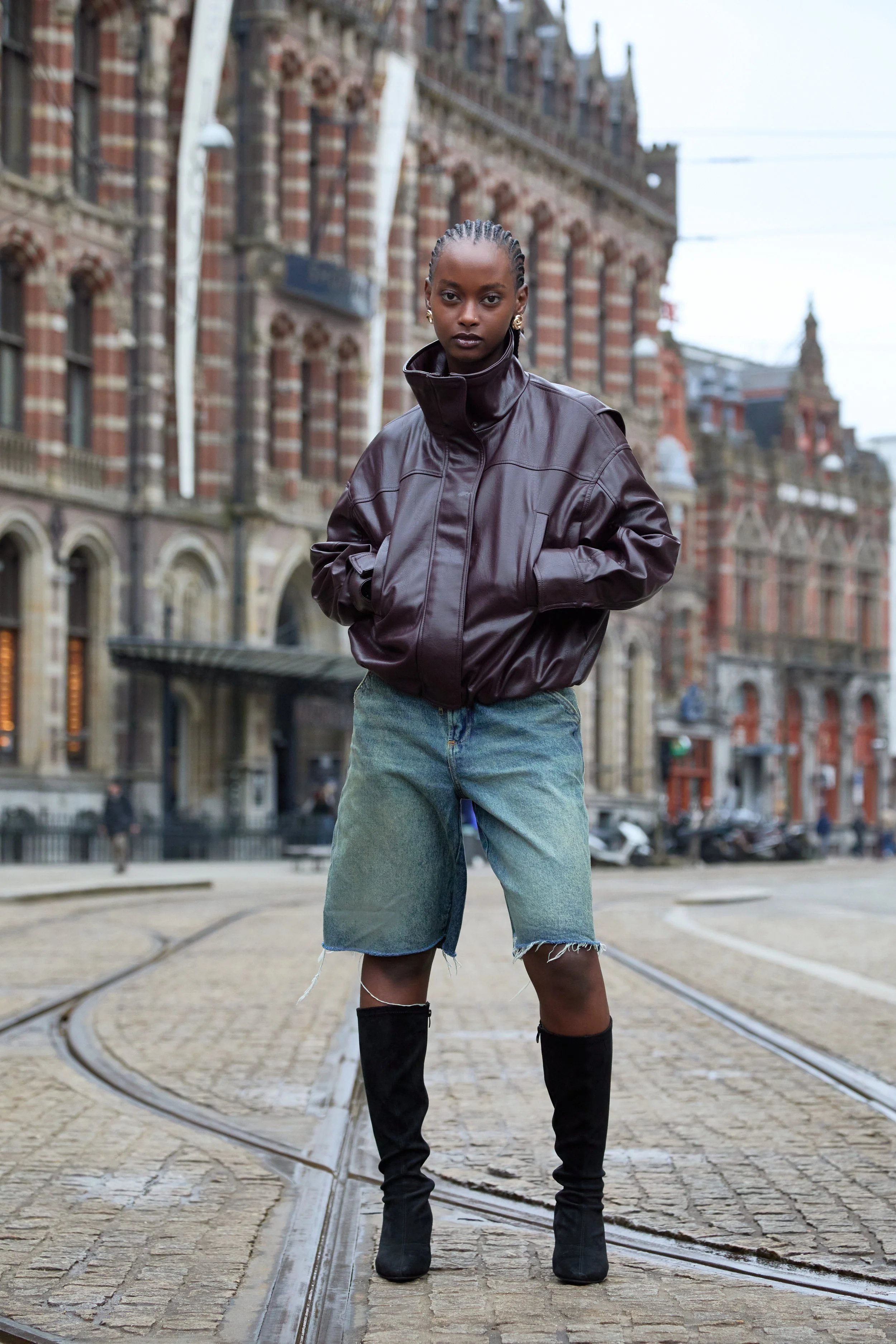 A woman standing on tram tracks in an urban setting, wearing a dark brown leather jacket, denim shorts, and black high-heeled boots.