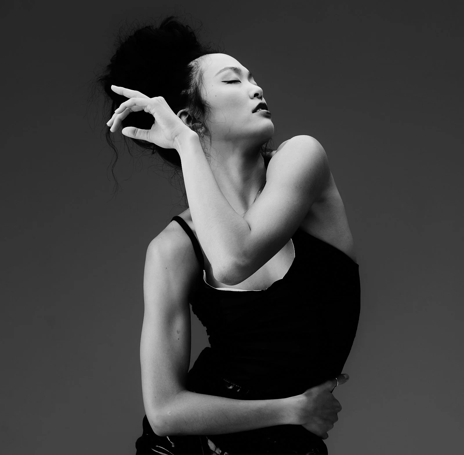 Black and white photo of a woman with her eyes closed, head tilted back, one arm crossing her body and the other hand near her face, wearing a black sleeveless top against a plain background.