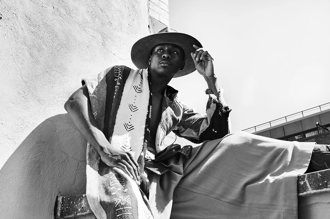 A person sitting outdoors in black and white. They are wearing wide-brimmed hat, loose pants, and a patterned jacket, posing against a textured wall with a bridge in the background.