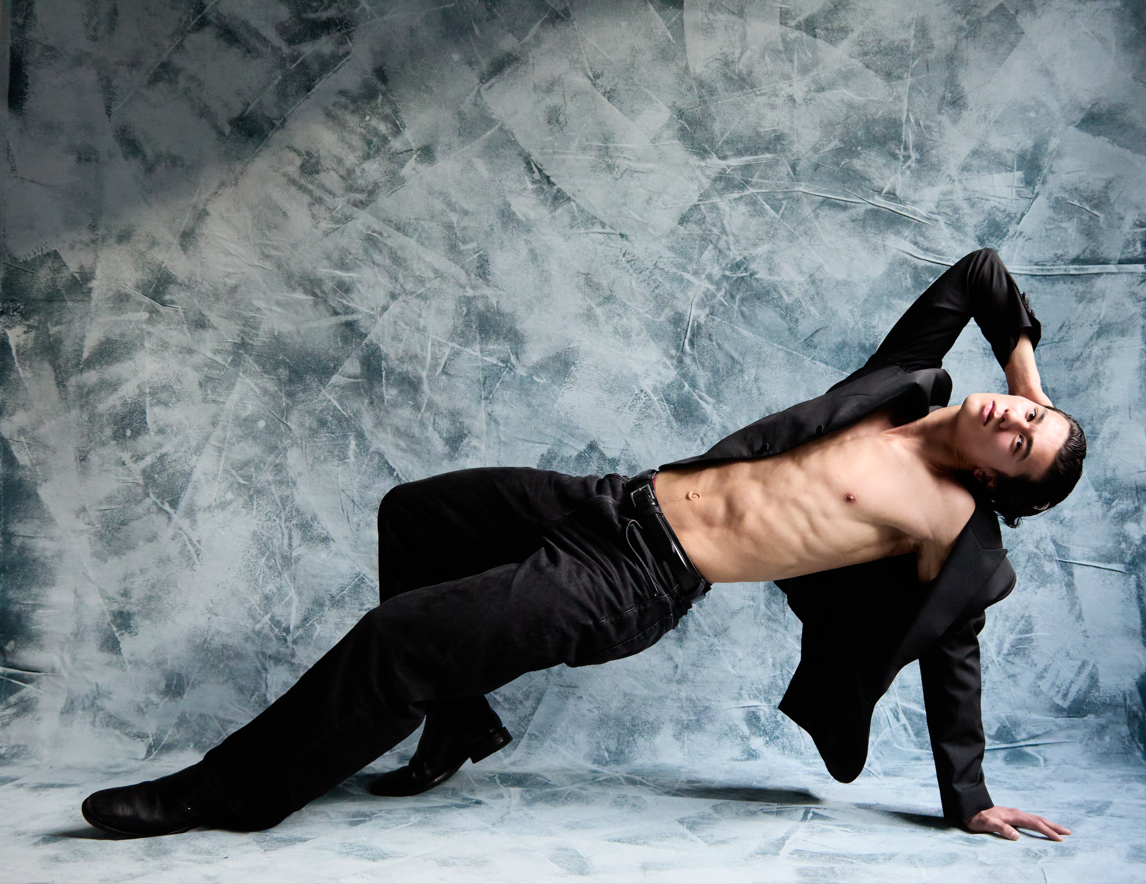 A shirtless man wearing black pants and shoes, posing in a contemporary studio with a textured gray wall background. He is leaning on one hand, with the other arm bent and resting on his head, showcasing his toned physique.