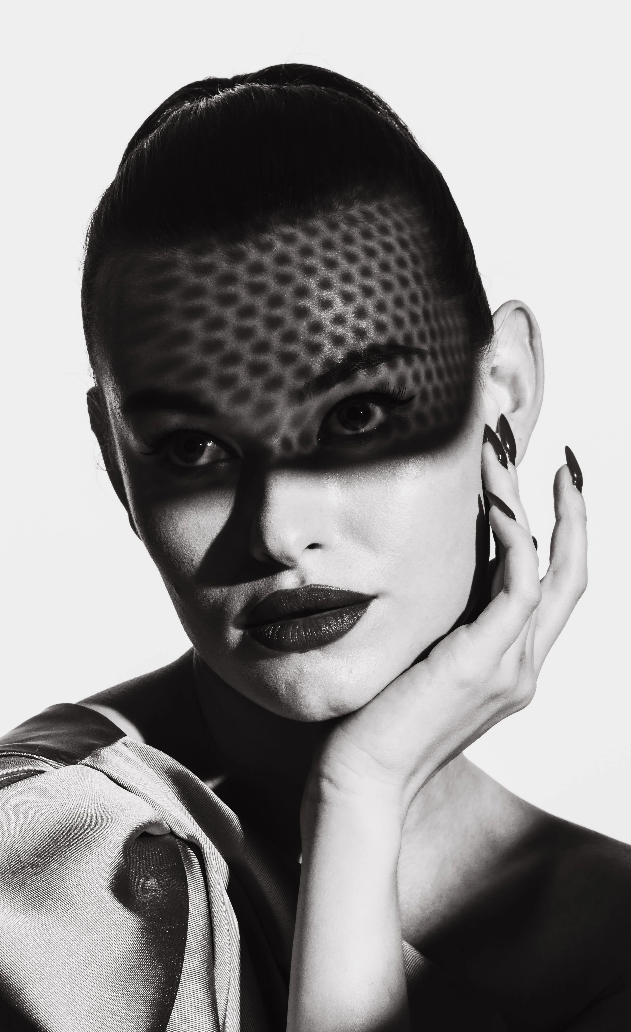 Black and white portrait of a woman with short dark hair, makeup, and dark lipstick, touching her face with her hand, wearing a shiny, textured garment.