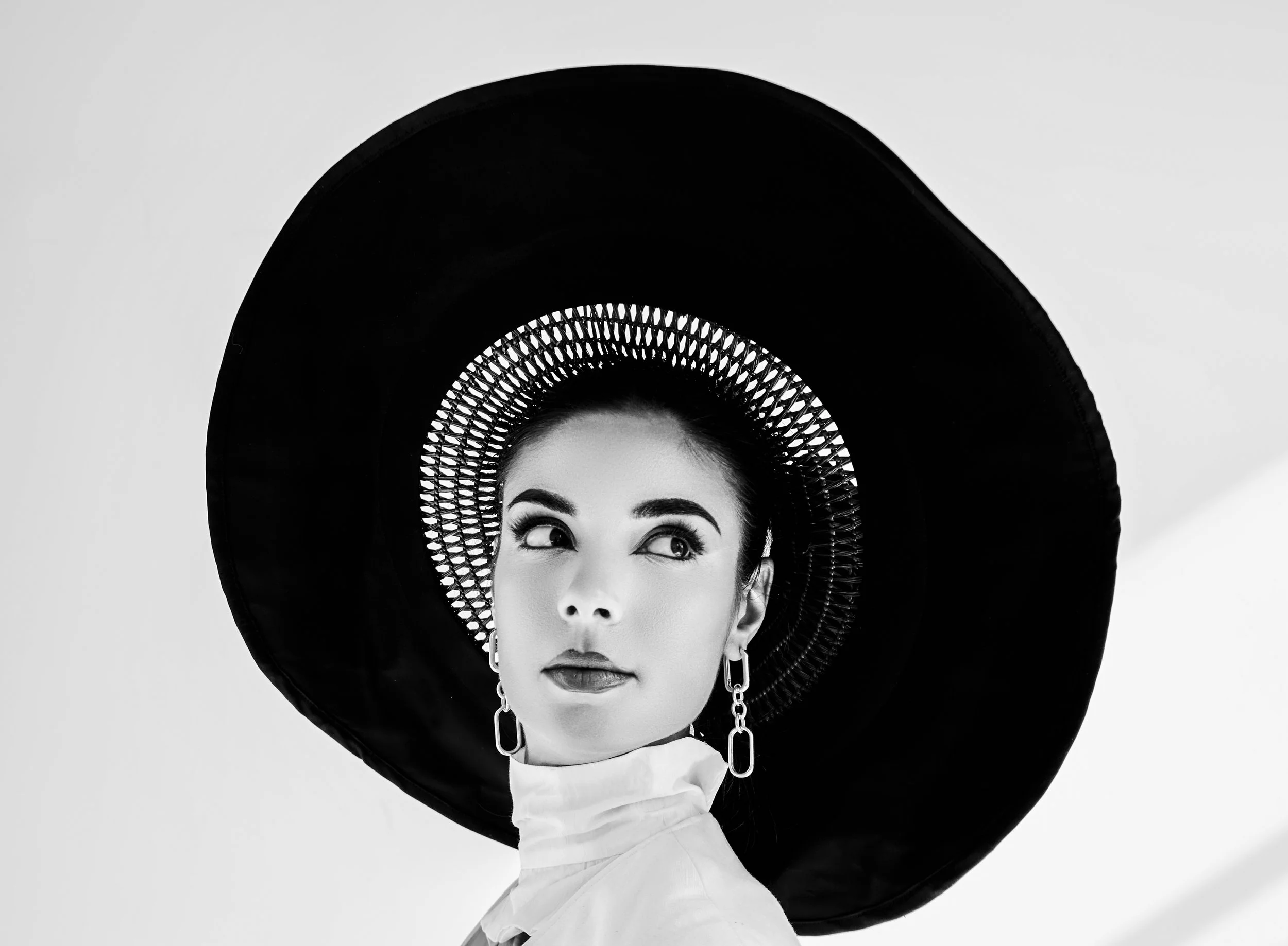 A black and white portrait of a woman wearing a large, wide-brimmed hat with intricate detailing around the crown, a high-collared shirt, and dangling earrings. She gazes off to the side with a composed expression.