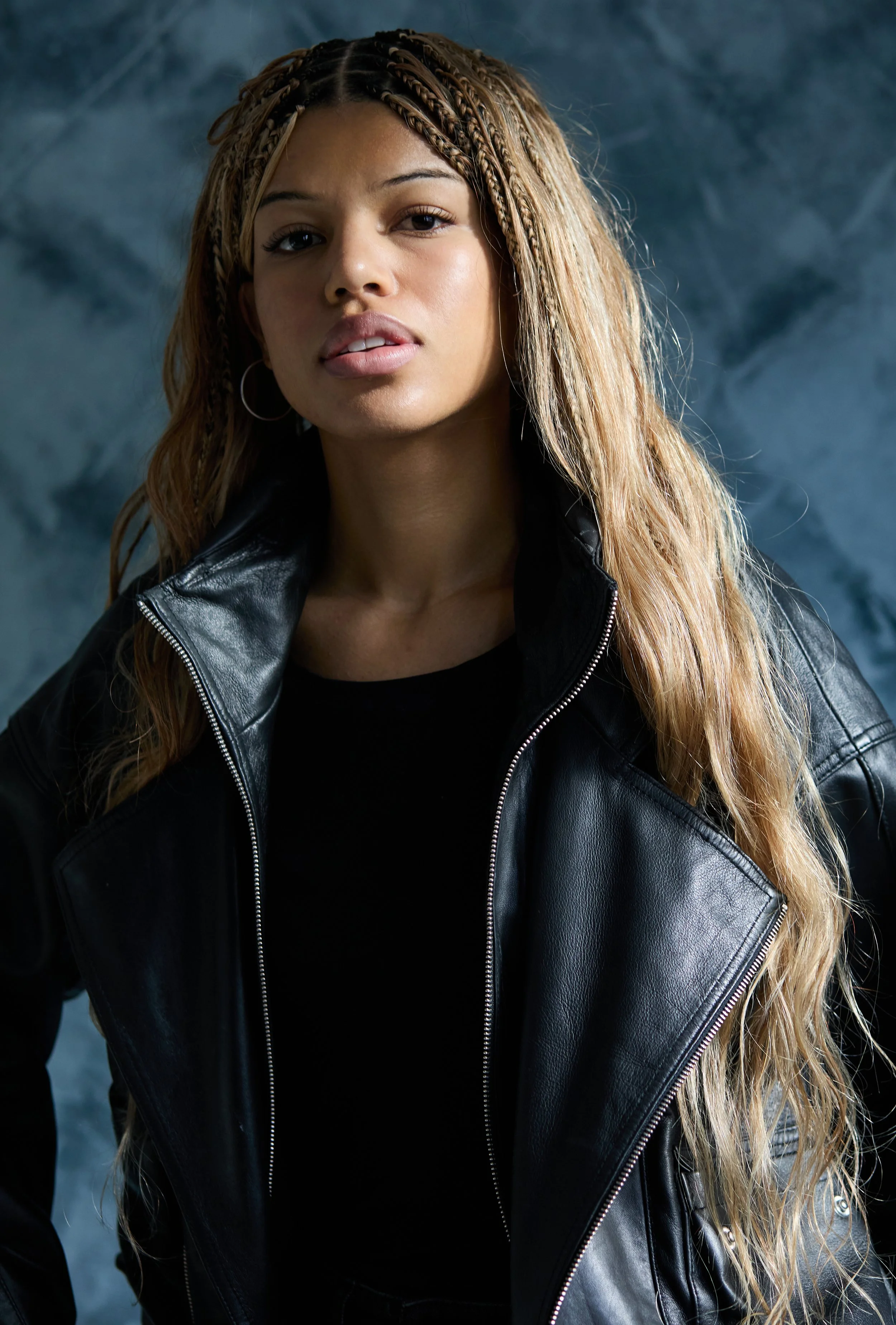 Portrait of a young woman with light brown braided hair, wearing hoop earrings and a black leather jacket, against a textured blue background.