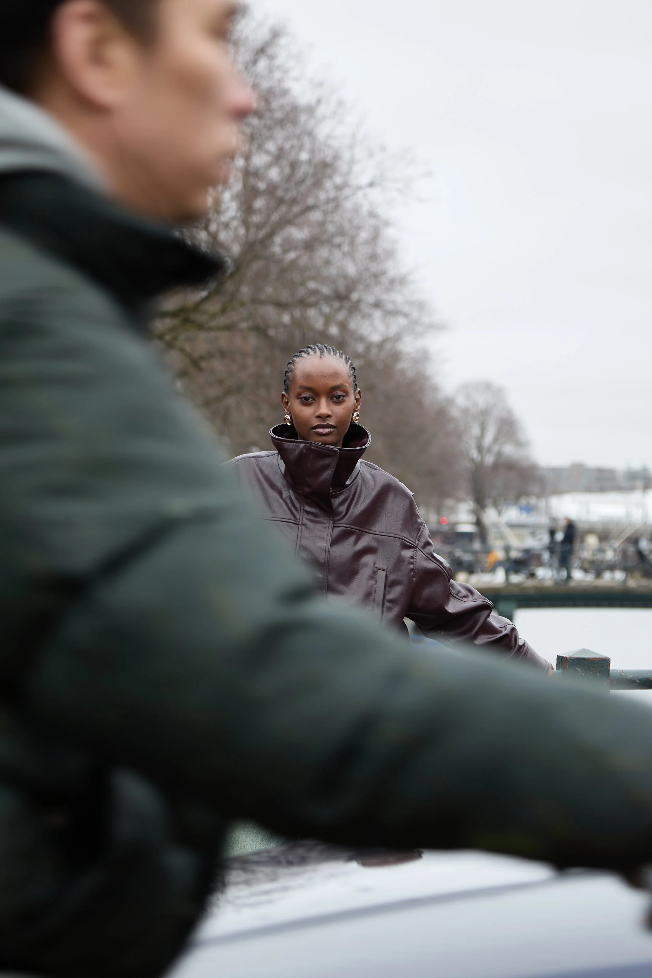 A woman with braided hair wearing a dark brown leather jacket, standing outdoors near a body of water with boats, trees, and an overcast sky in the background, while a person in a gray jacket is partially visible in the foreground.