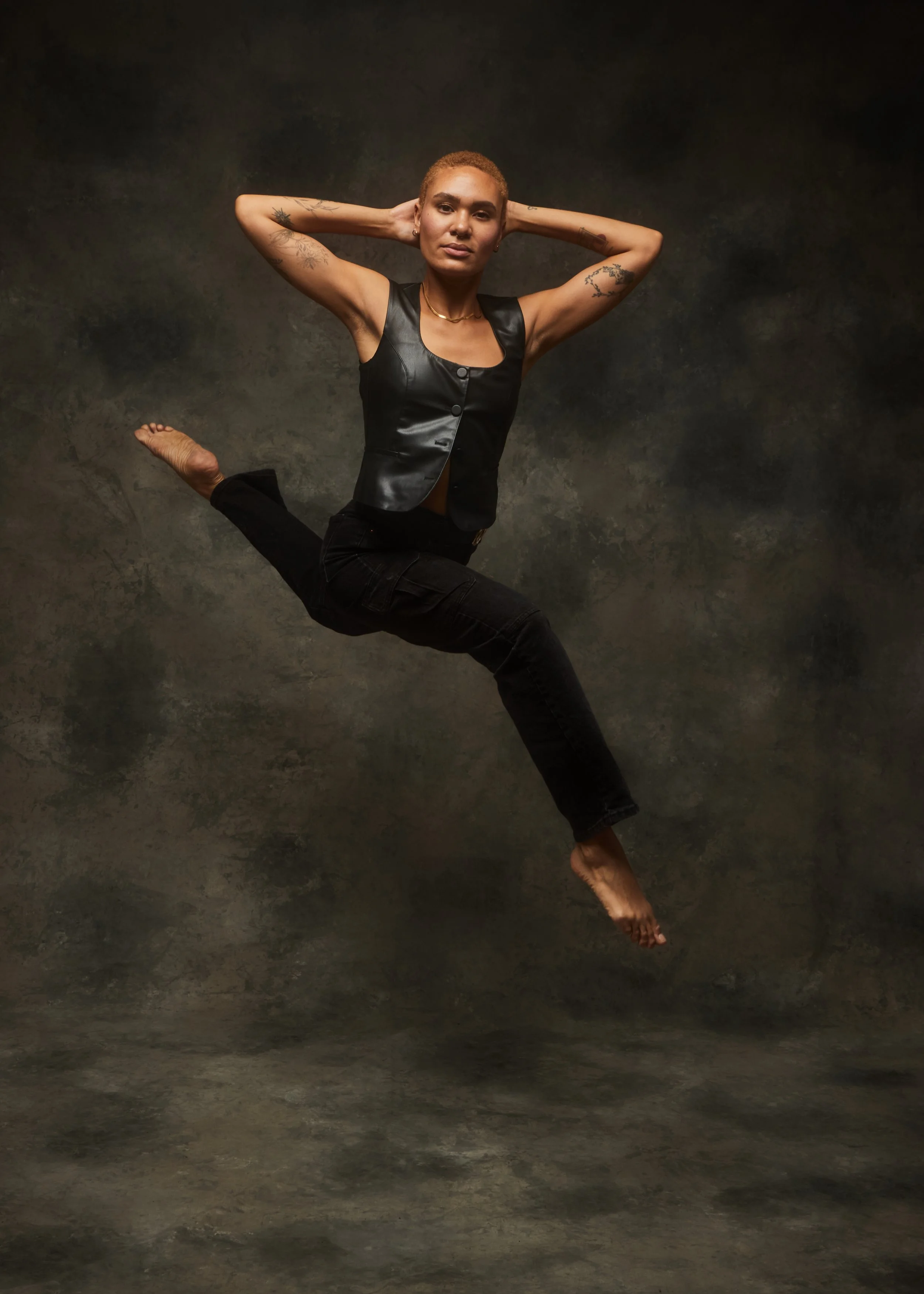 A woman with short hair and tattoos, wearing a black sleeveless top and black pants, is posing against a dark textured background with her arms behind her head and one leg lifted.