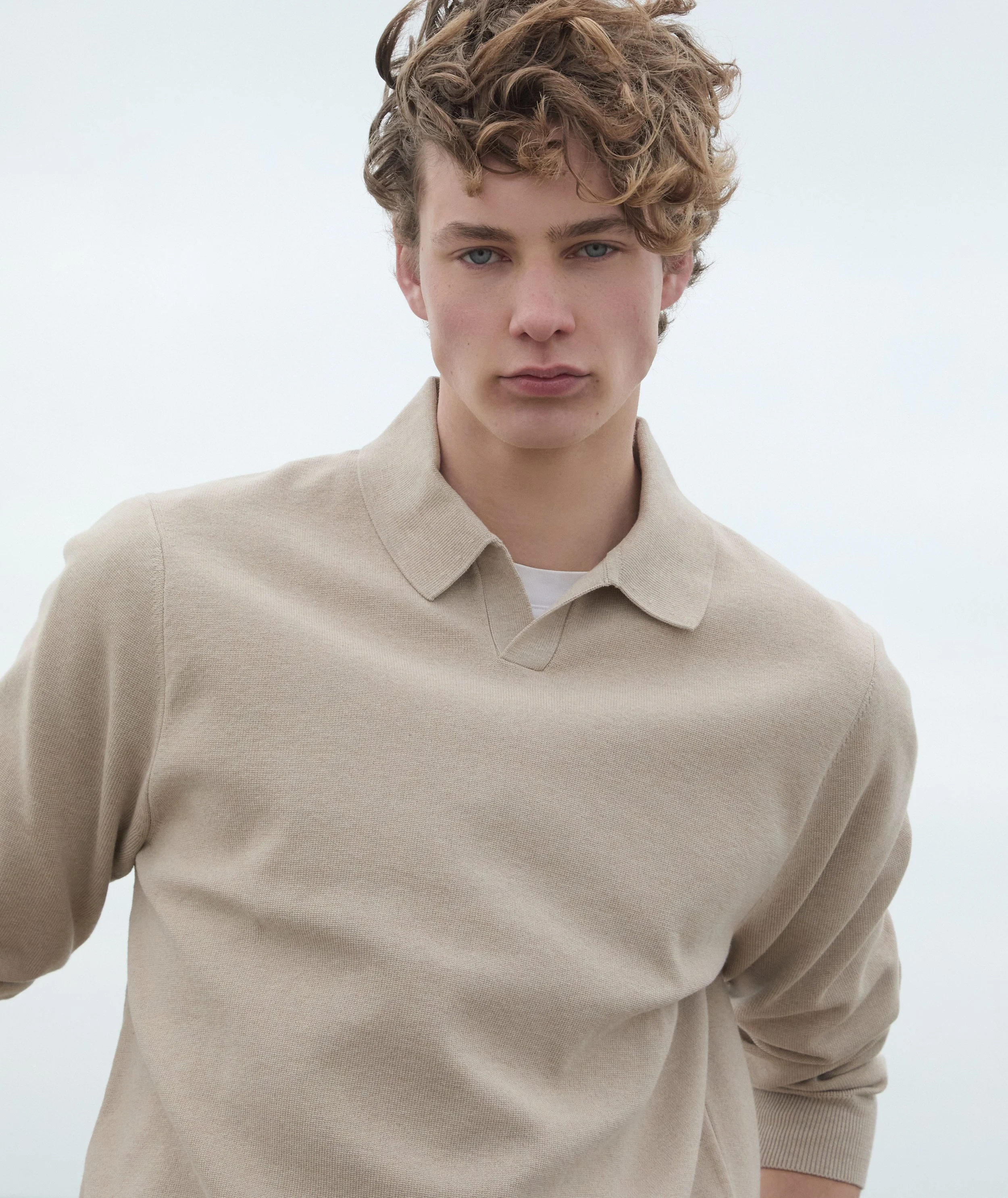 Young man with curly blonde hair wearing a beige collared shirt, standing against a cloudy sky.