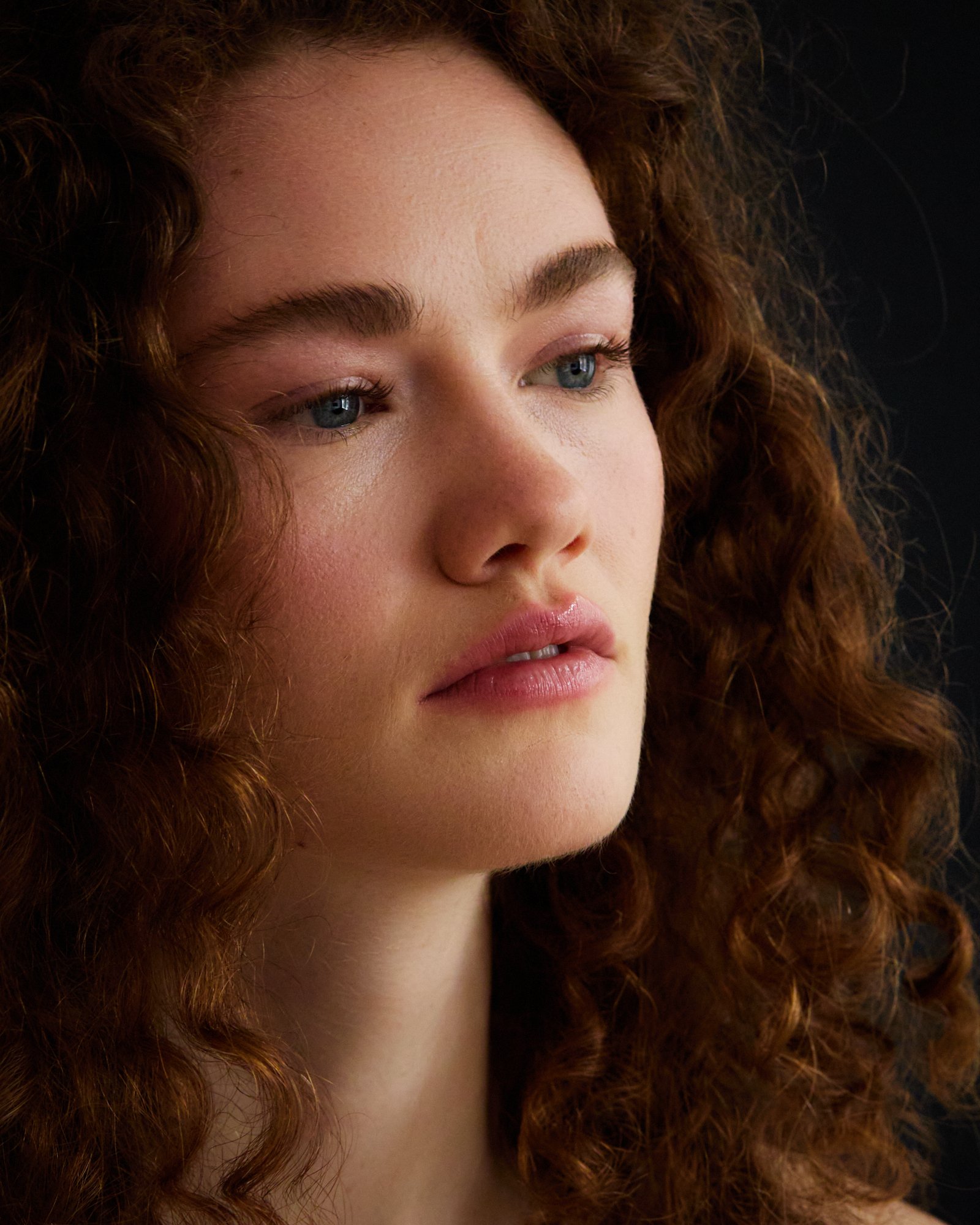 Close-up of a woman with red, curly hair and blue eyes, looking to the right against a dark background.