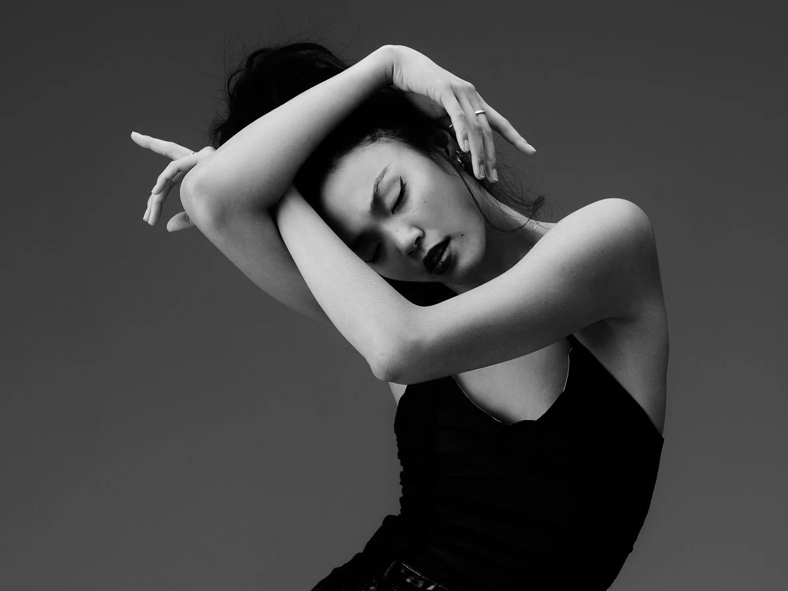 A woman with dark hair posed with her arms crossed over her face, wearing a black sleeveless top, against a solid dark background in a black-and-white photograph.