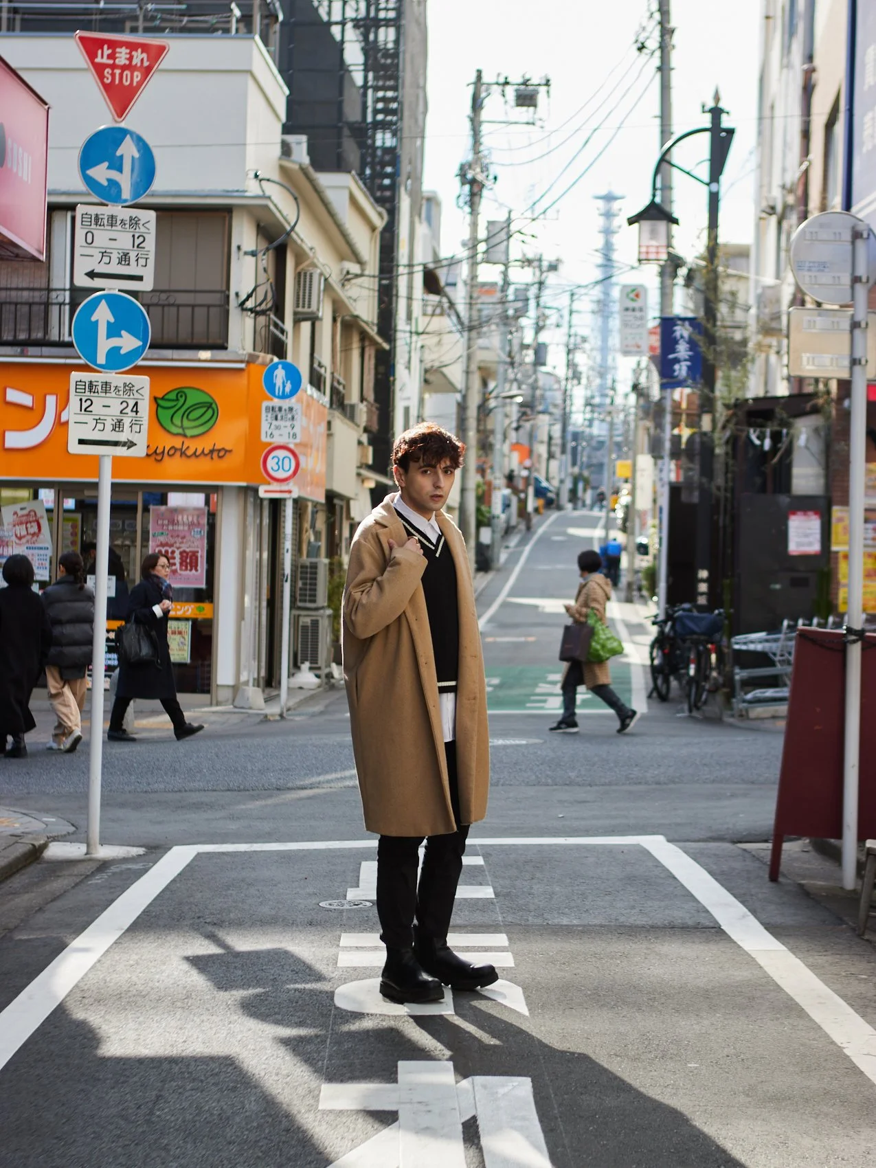 A young man in a tan coat standing on a pedestrian crossing in an urban street, with shops, pedestrians, and power lines visible in the background.