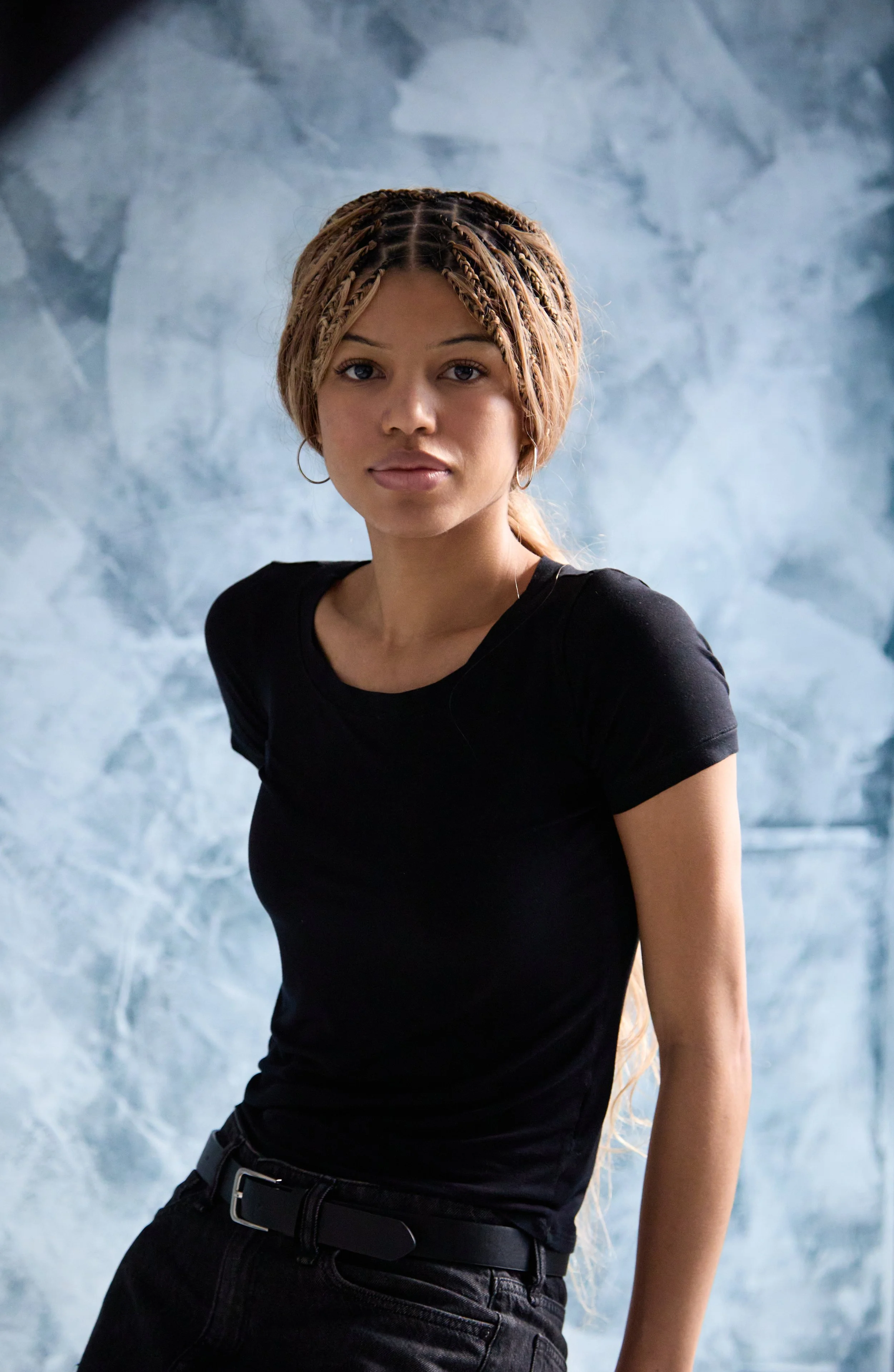 Portrait of a young woman with braided hair, wearing a black t-shirt and black pants, standing in front of a textured blue-gray background.