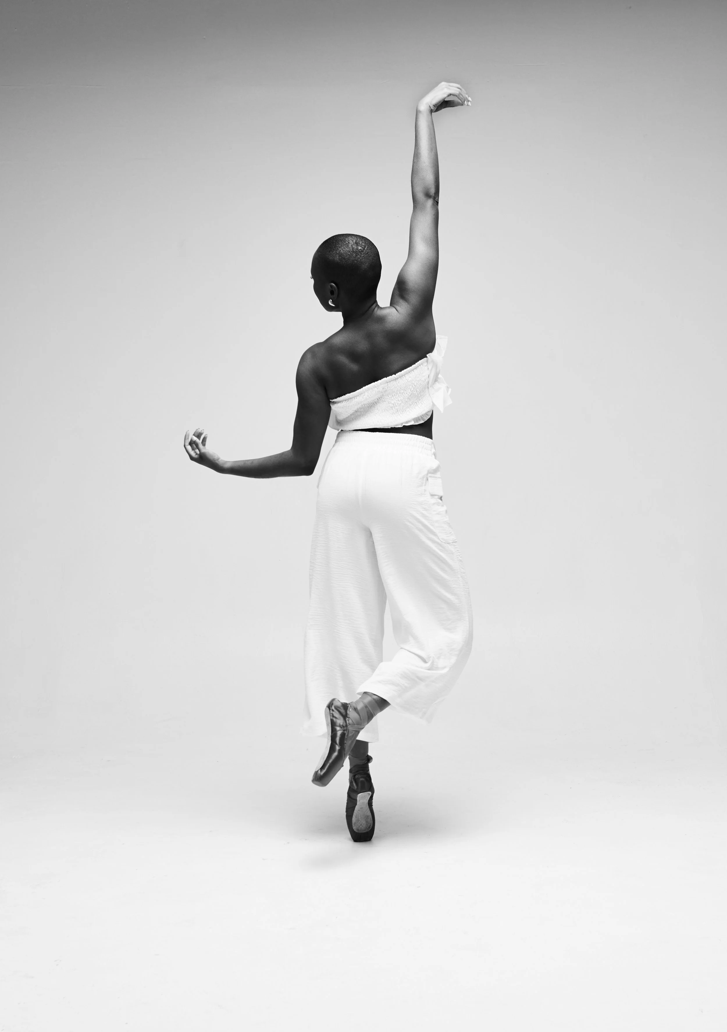 Black and white photo of a ballet dancer mid-performance, balancing on one toe with arms gracefully extended, dressed in a strapless top and wide-leg pants.