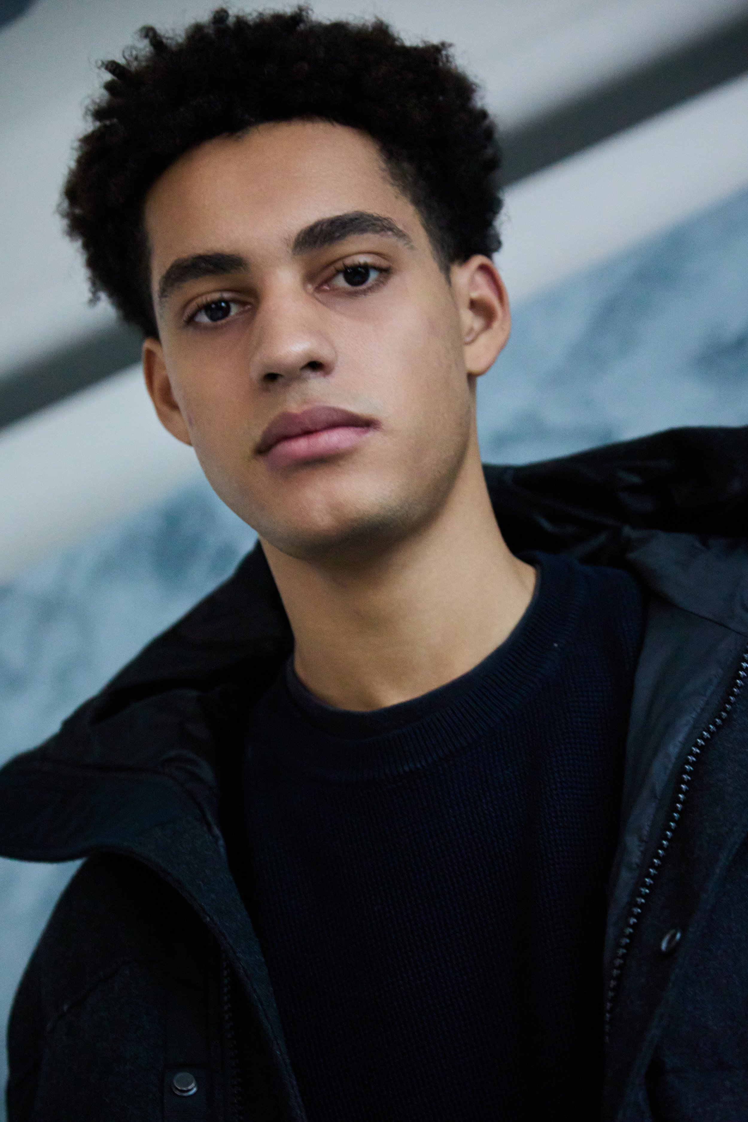 A young man with short curly dark hair, wearing a black jacket over a black shirt, looking directly at the camera with a neutral expression. The background is blurred but appears to be indoors.