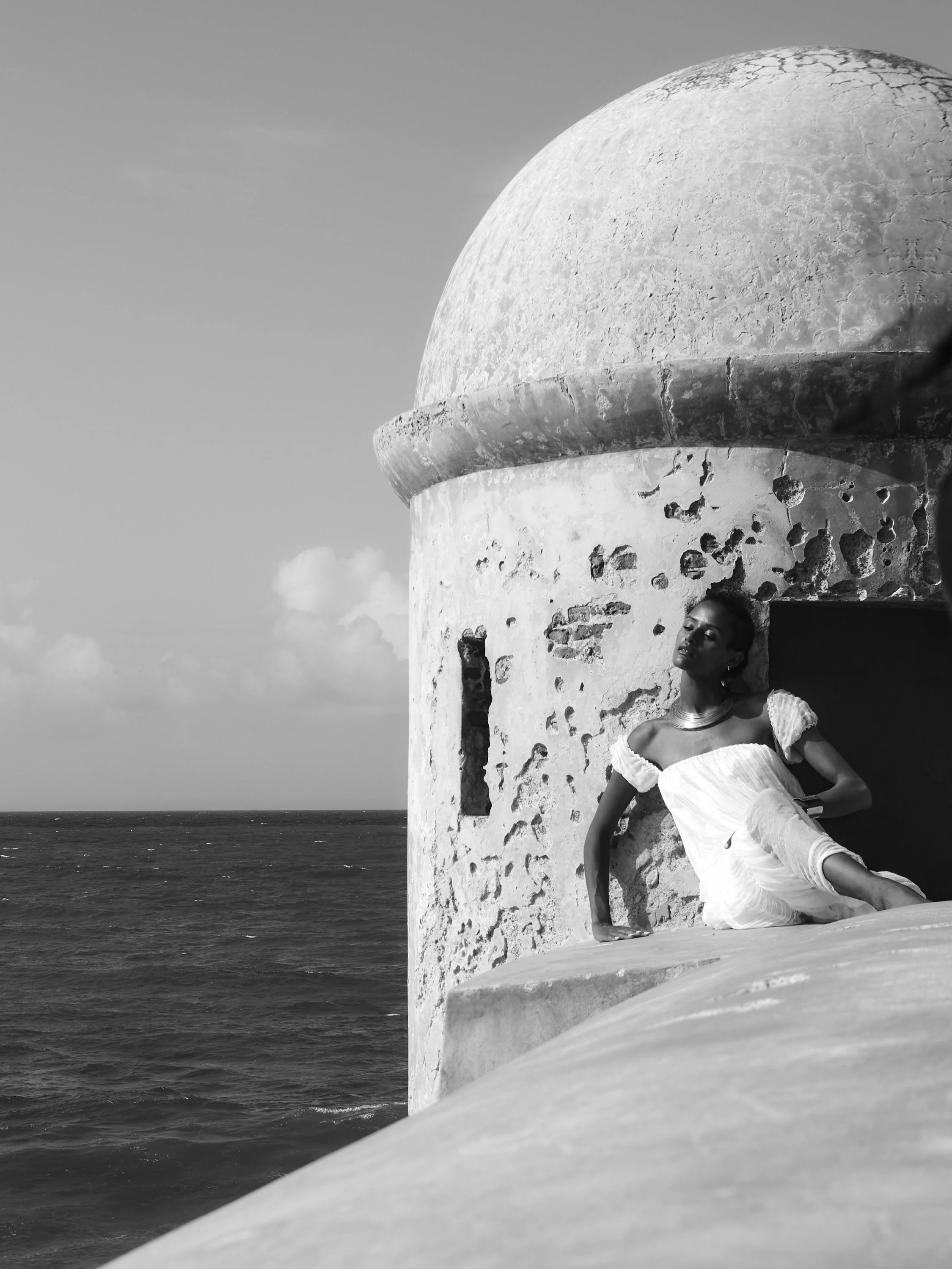 A woman in a white dress with ruffled sleeves and a necklace sitting on a ledge outside a cylindrical, textured, domed building by the sea in black and white.