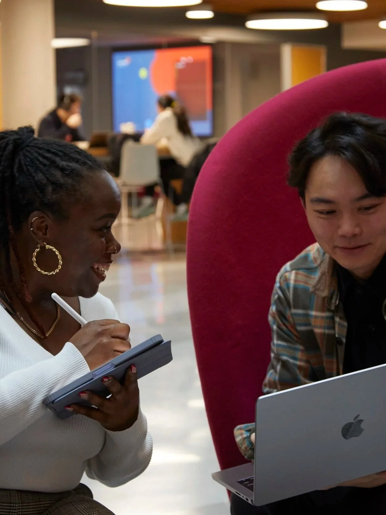 Two people sitting together in a modern office or lounge, looking at a laptop and a tablet, engaged in conversation. In the background, other individuals are working at a table with a large digital screen on the wall.