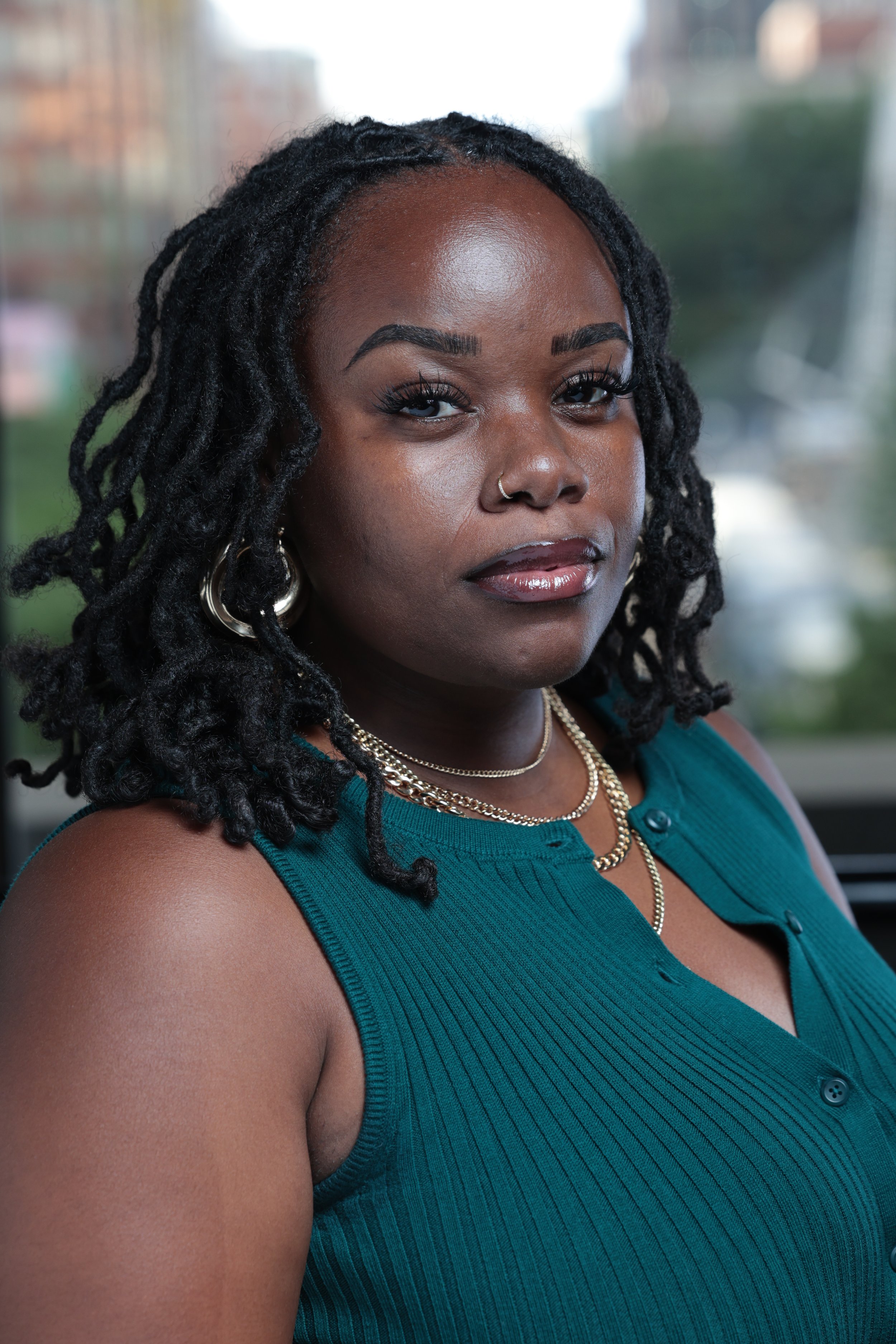 A Black woman with dreadlocks, wearing a teal sleeveless top, gold necklaces, hoop earrings, and a nose ring, looking confidently at the camera with a blurred cityscape background.