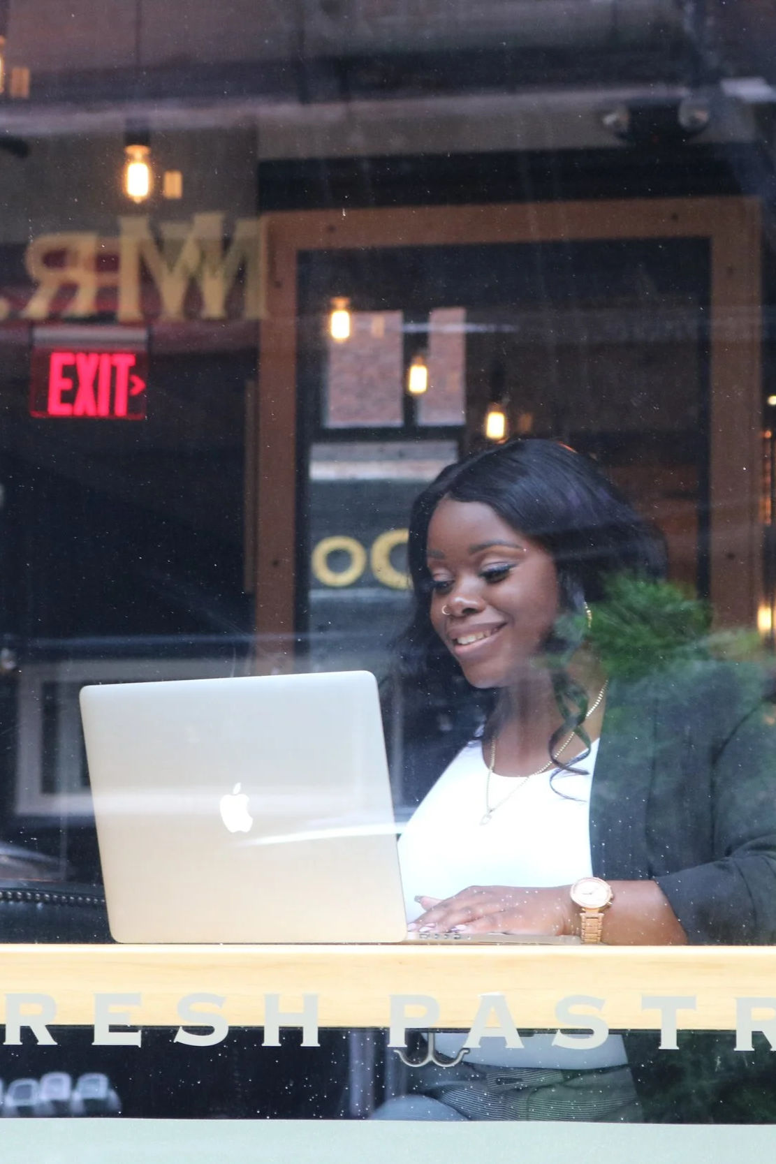A woman with black hair working on a silver Apple MacBook laptop in a coffee shop seen through a window.