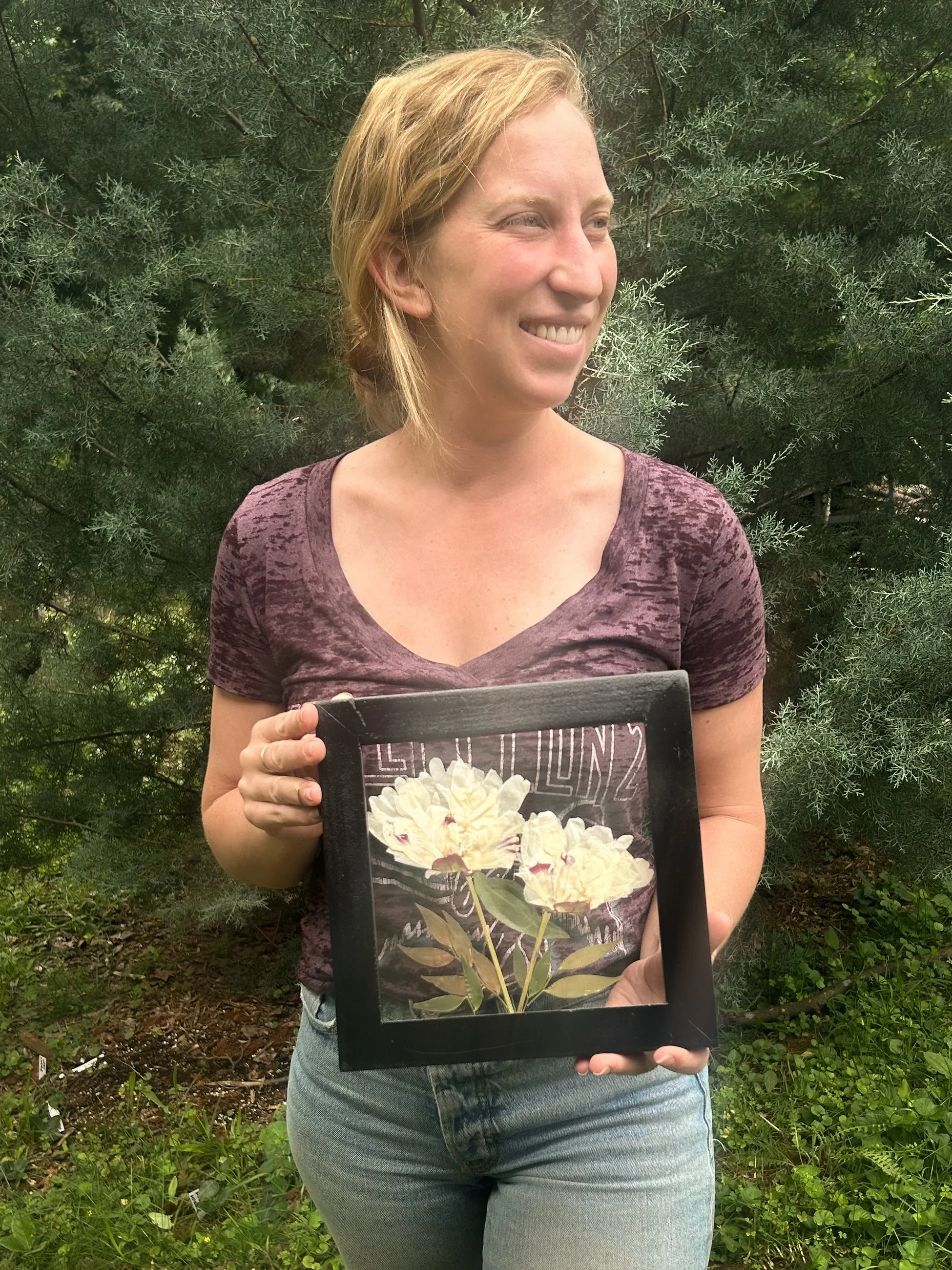 A woman with blonde hair smiling while holding a flower picture frame in front of her, outdoors with green foliage in the background.