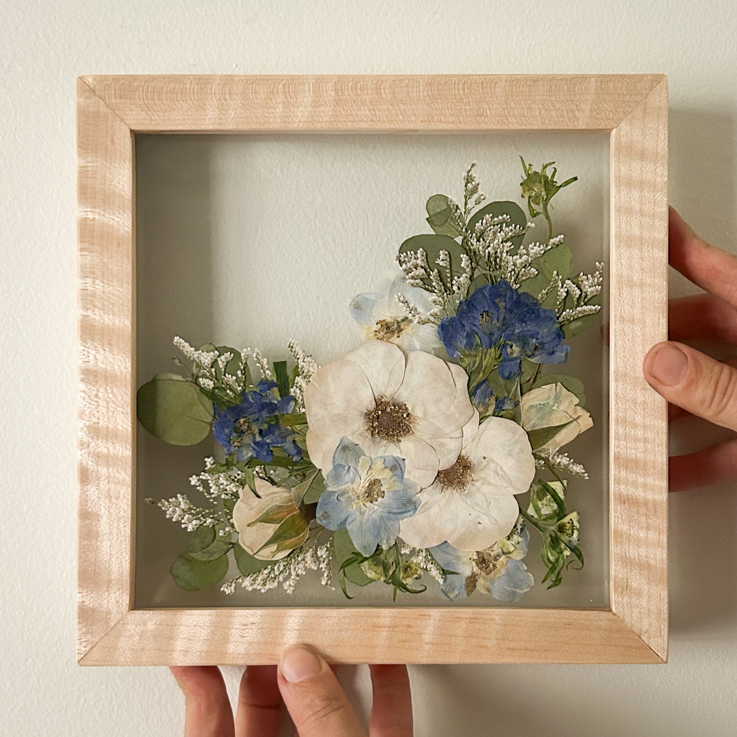 A person holding a framed picture of white flowers and green leaves with a wooden background.