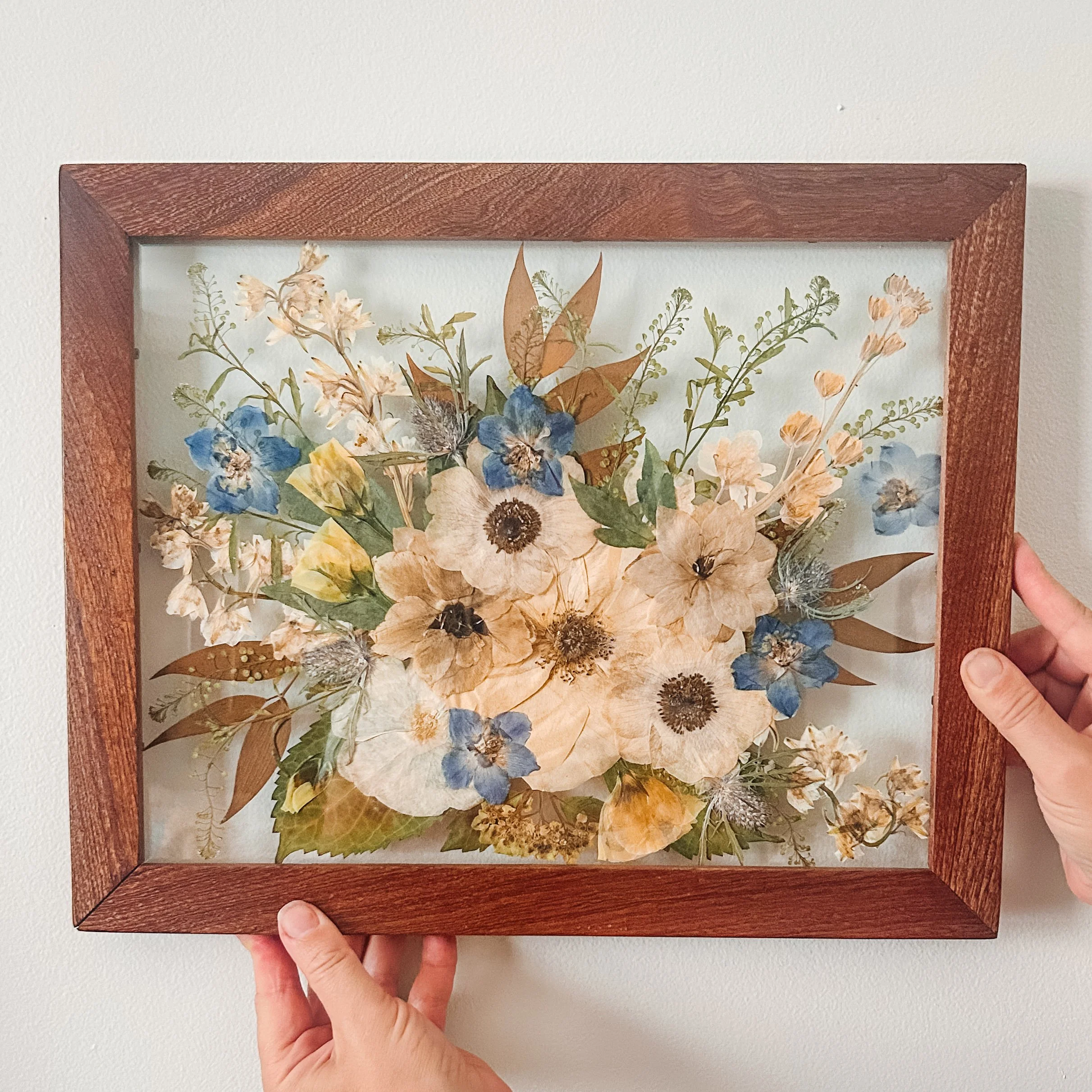 A hand holding a framed botanical print featuring dried flowers and leaves against a white wall.