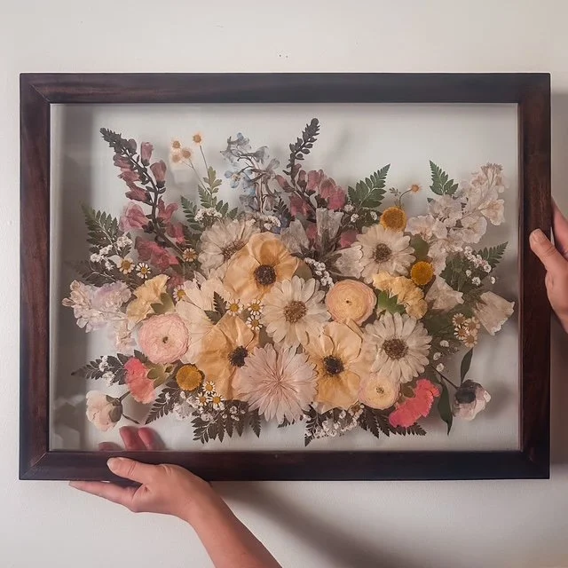 Framed pressed flower art featuring assorted dried flowers and greenery displayed on a white wall.