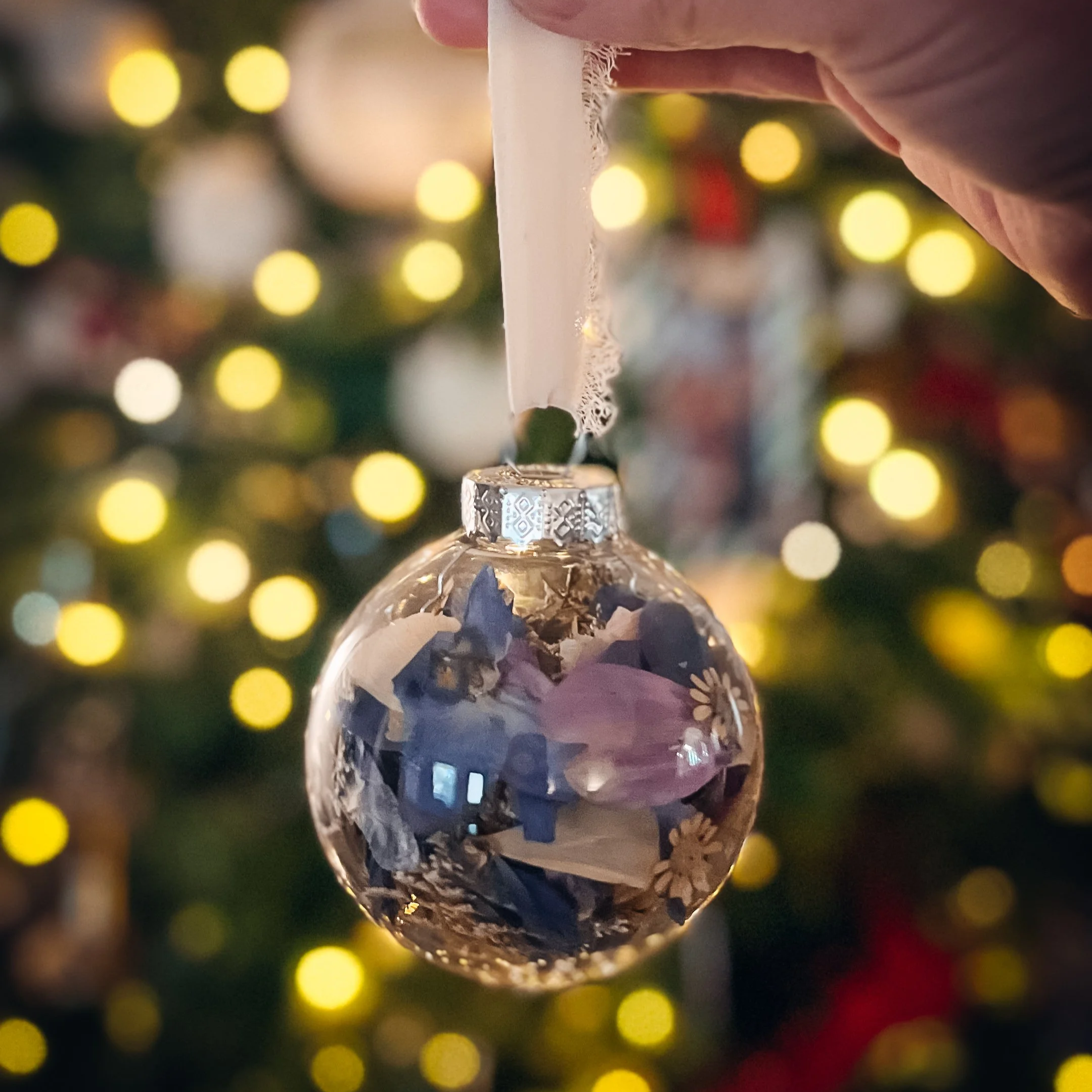 A glass Christmas ornament with a reflective surface hanging from a Christmas tree. The background features warm yellow bokeh lights.