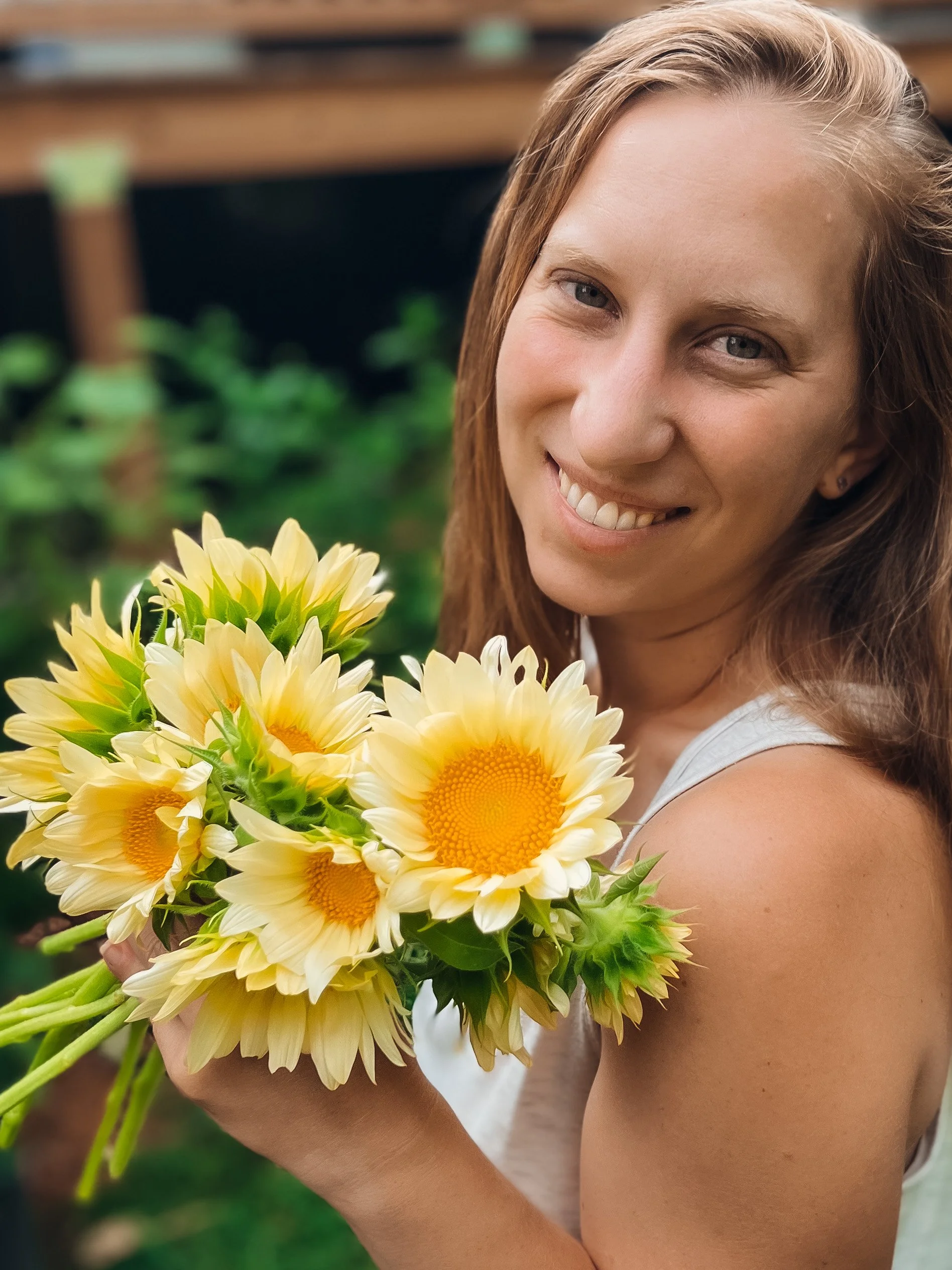 A woman with long brown hair and a white sleeveless top holding a bouquet of yellow and white daisies, smiling in a garden setting.