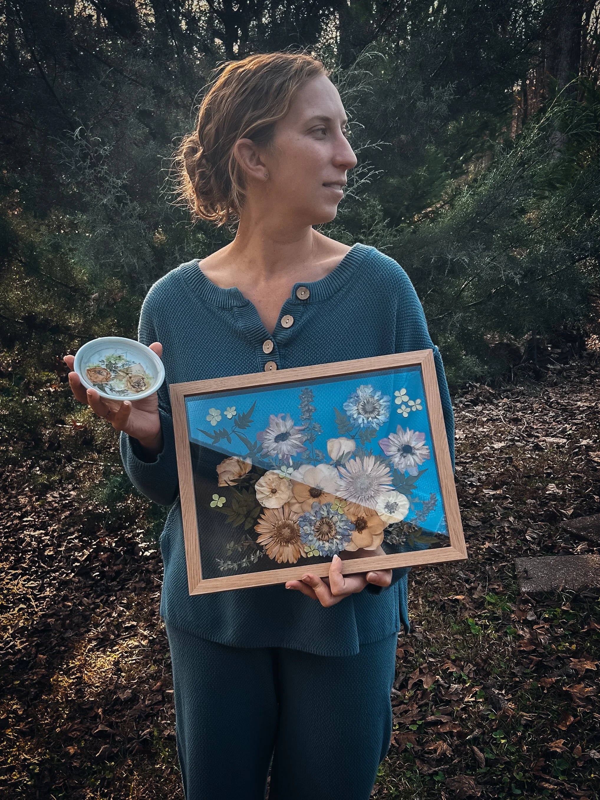 A woman with short blond hair in a braid, wearing a blue sweater, standing outdoors among trees and holding a framed floral artwork and a small dish with dried flowers.