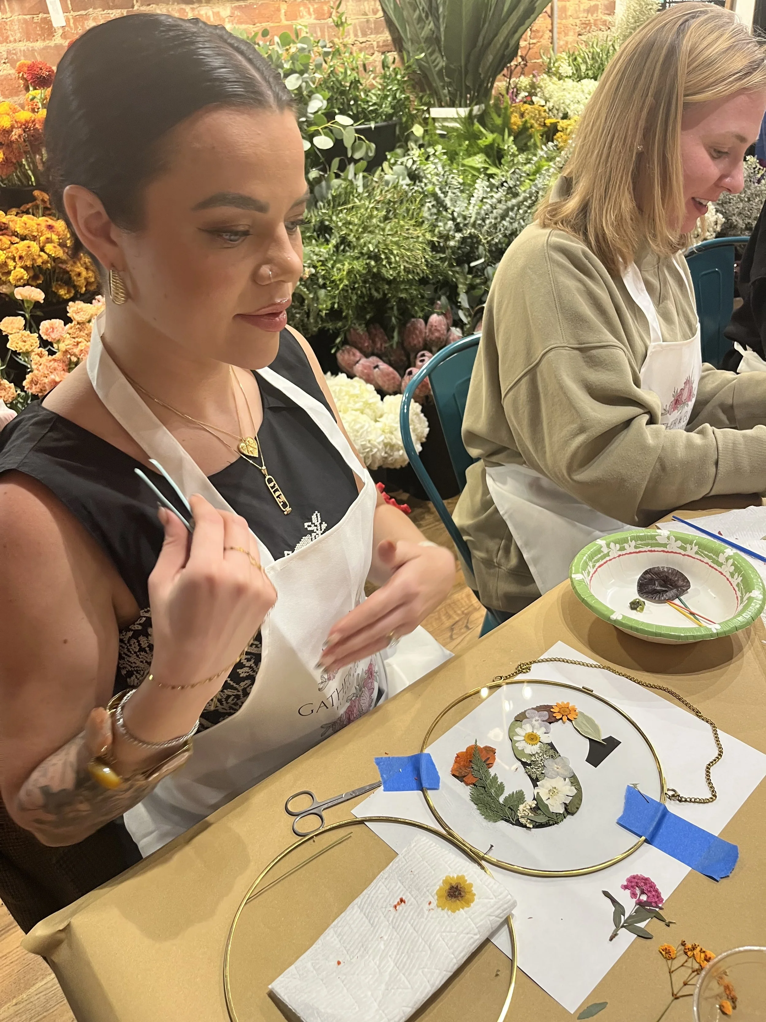 Two women sit at a table working on a craft project involving pressed flowers and leaves. The table has various crafting supplies, including scissors, tape, paper towels, and a decorative wreath with pressed flowers. Behind them are colorful flowers and green plants indoors.