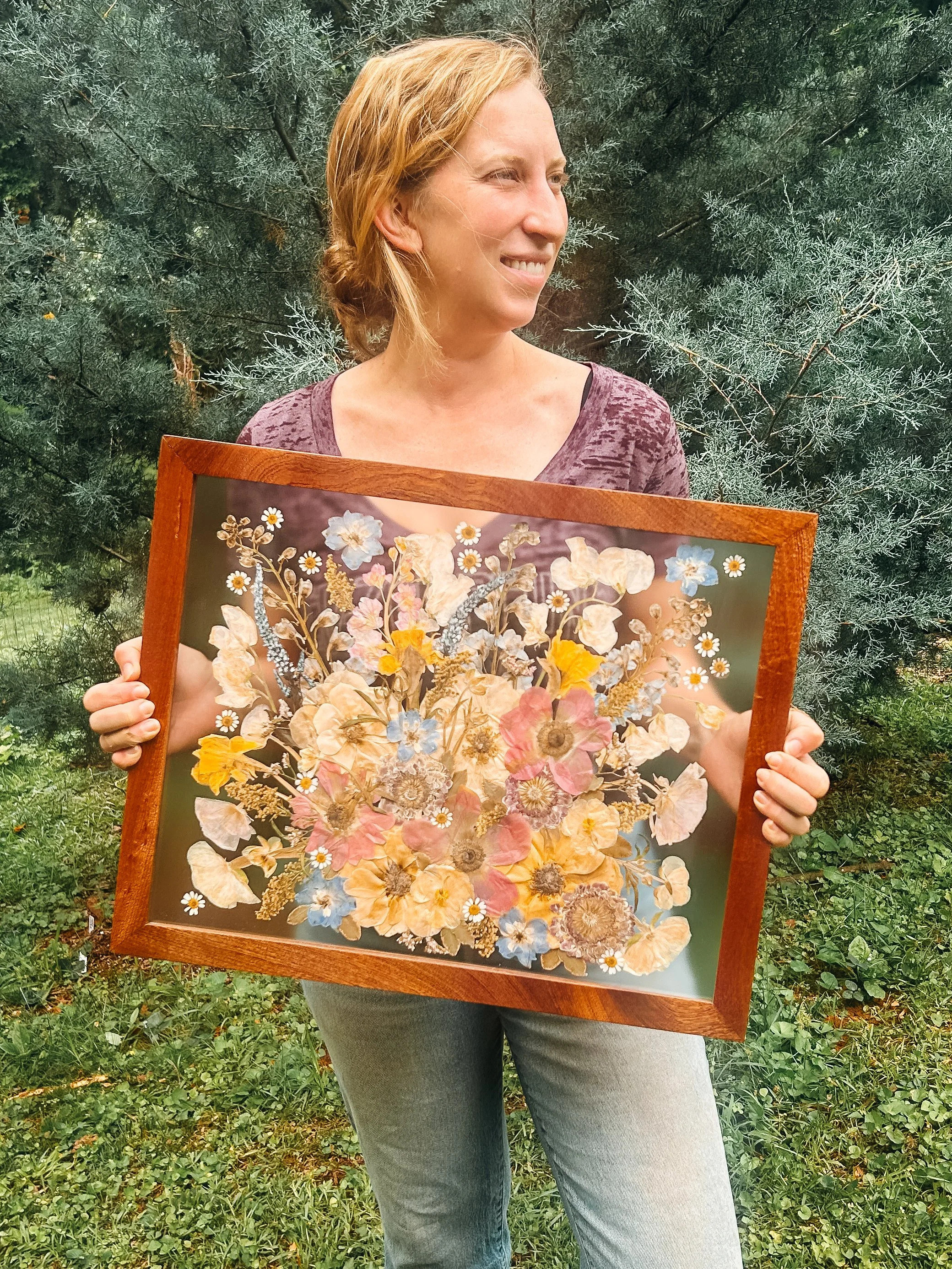 A woman with short blonde hair smiling and holding a framed picture of dried flowers outdoors in front of green trees and grass.