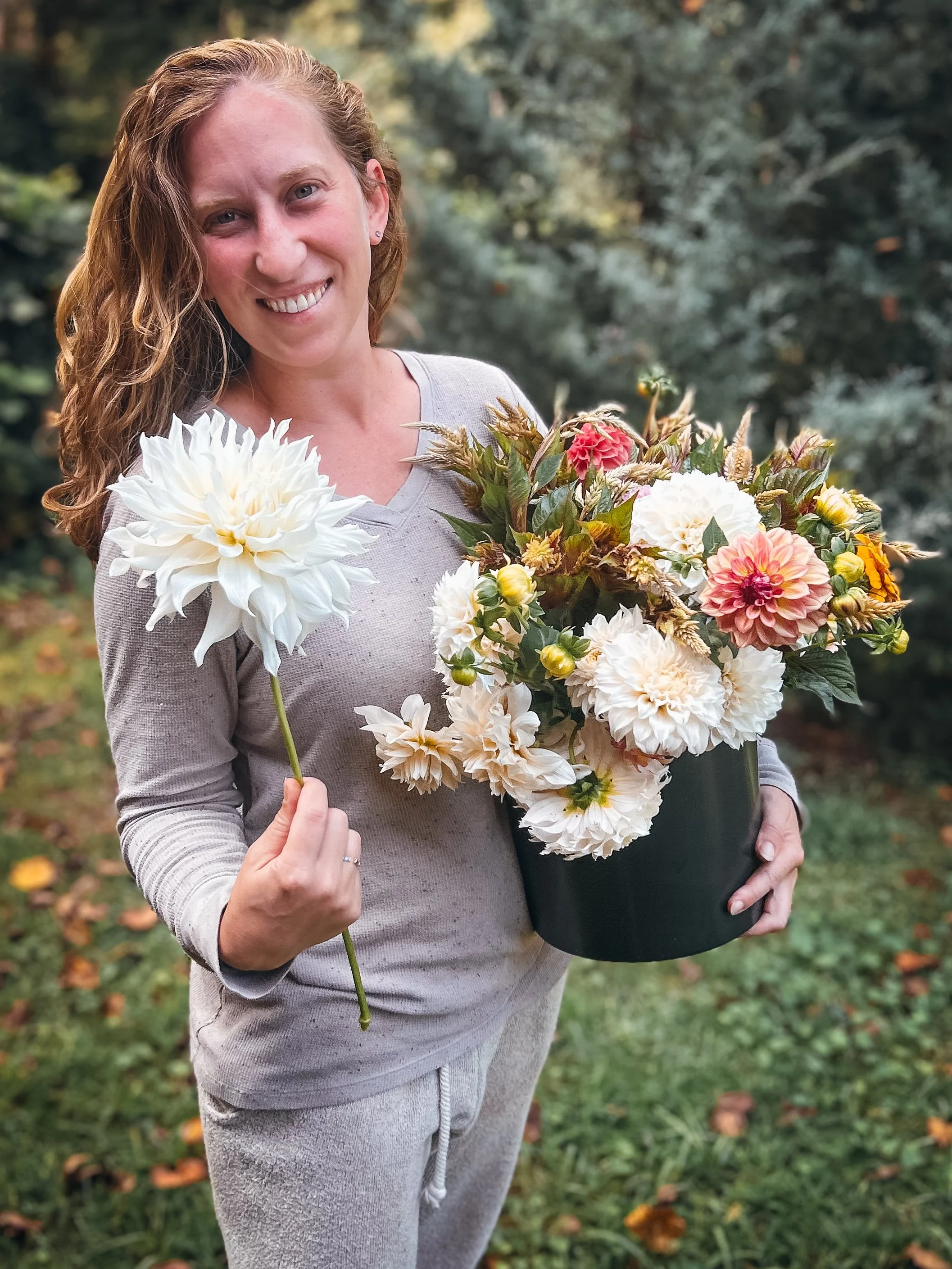 A woman holding a large black pot filled with various flowers, including white dahlias and pink and red blossoms, outdoors in a garden with a blurred background of trees and grass.