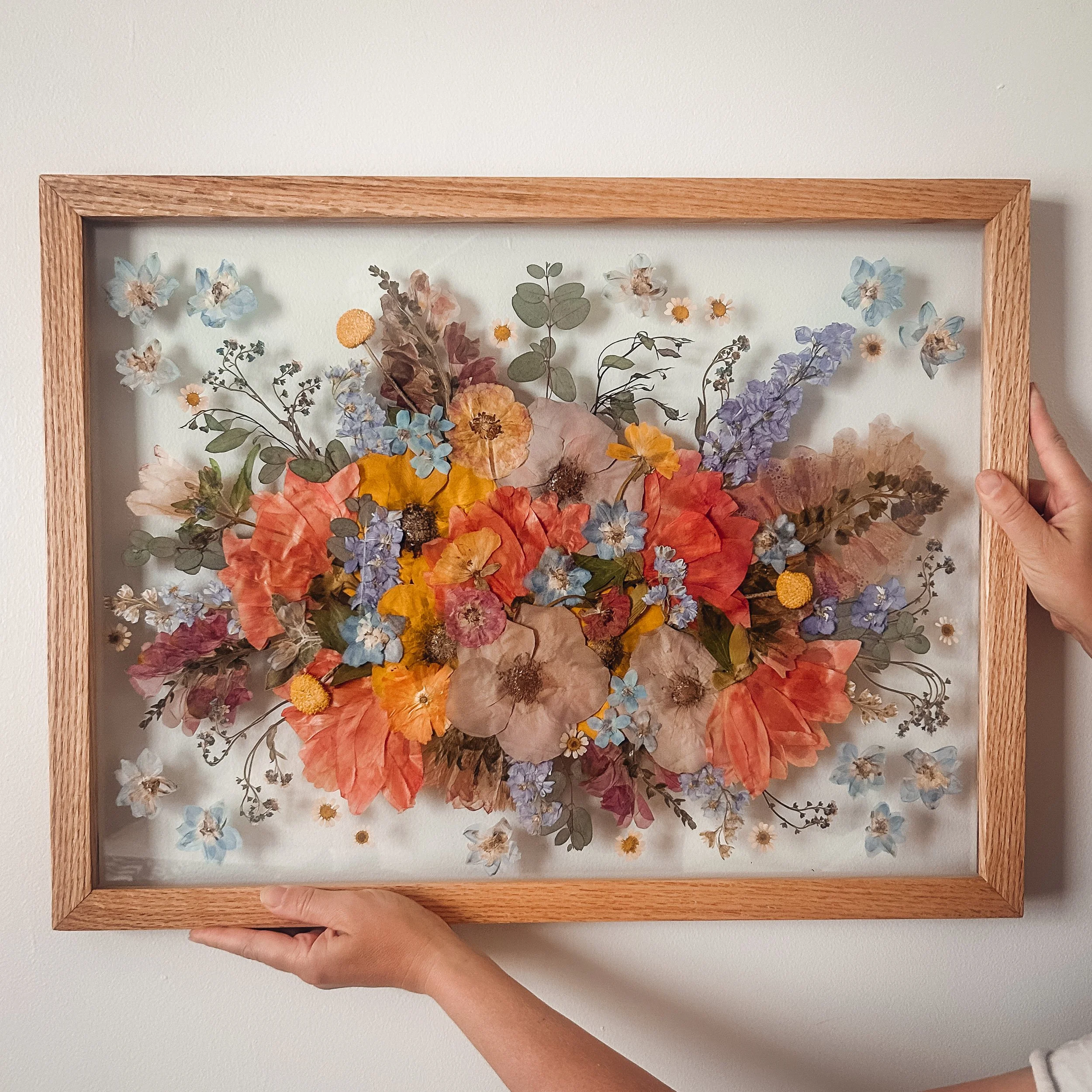 A person holding a framed pressed flower collage against a plain white wall.