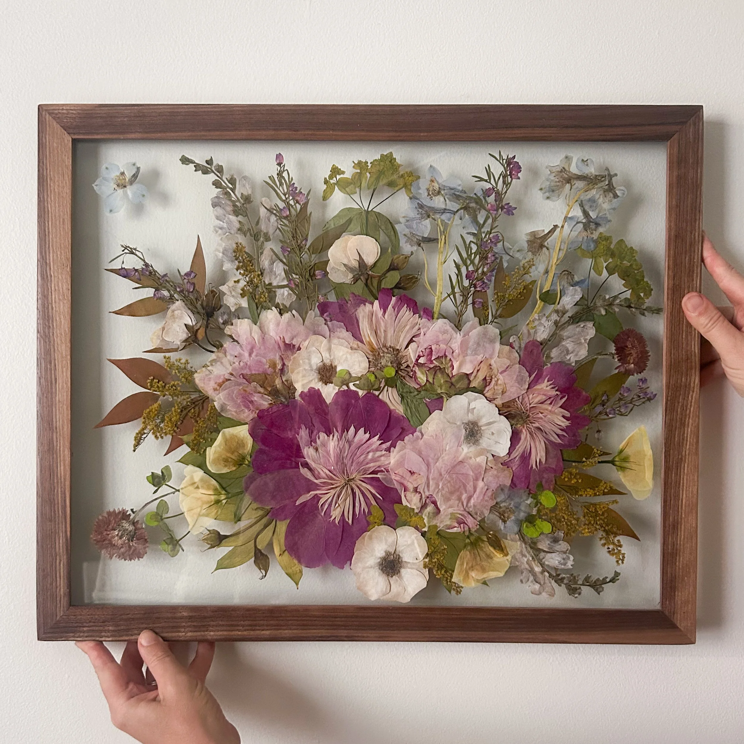 Person holding a framed artwork of dried and pressed flowers against a green leafy background. The person wears a sleeveless top with black and white stripes.