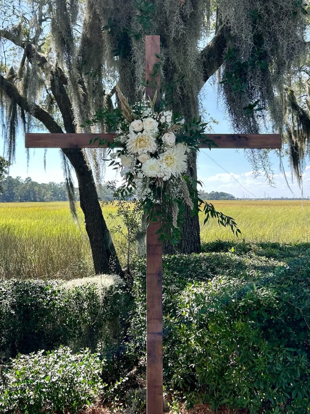 Wooden cross decorated with white flowers, greenery, and baby’s breath stands outdoors in a grassy field with trees and a clear blue sky in the background.