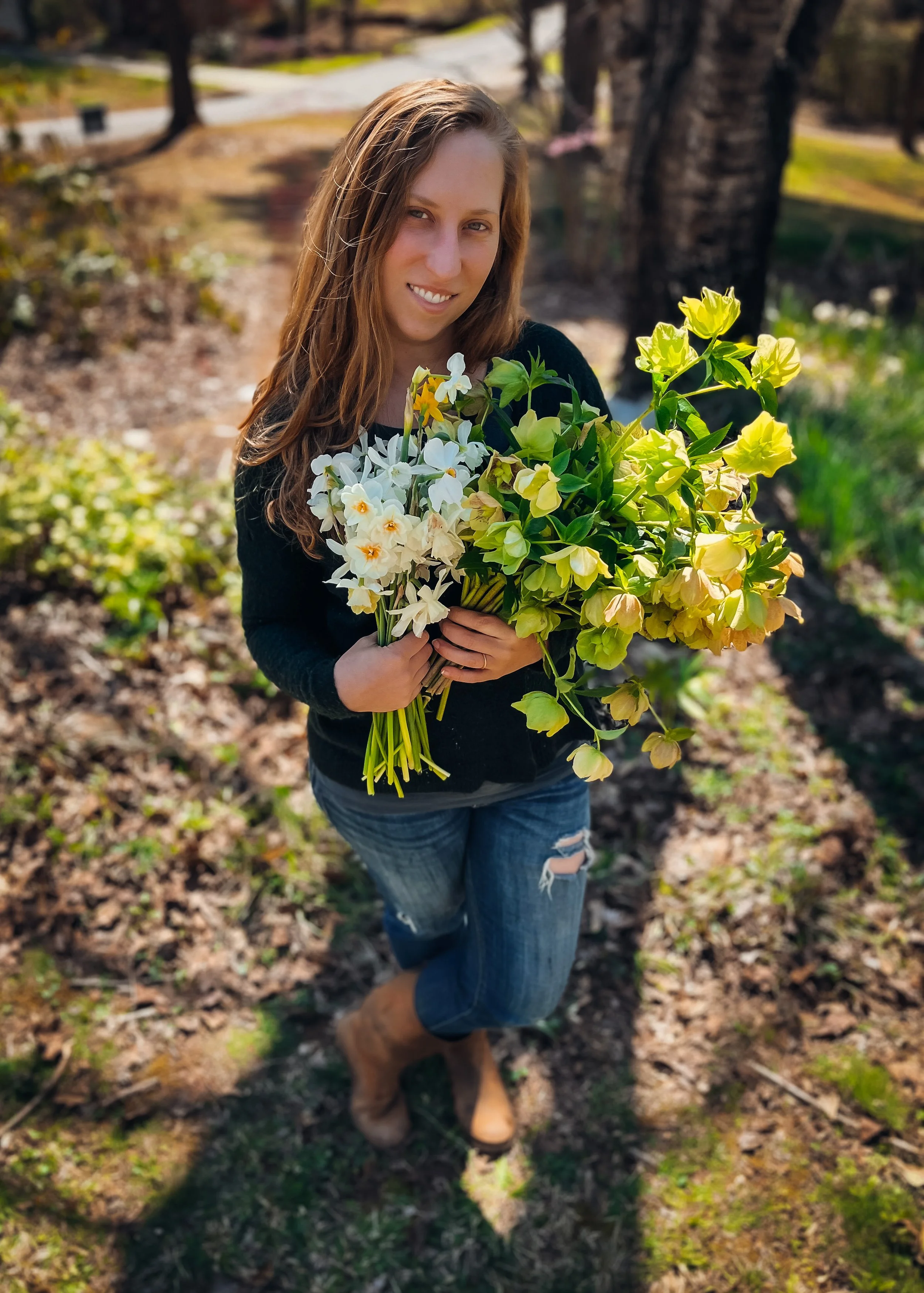 A woman with long, wavy brown hair smiling while holding a large bouquet of yellow and white flowers in an outdoor park with trees and fallen leaves.