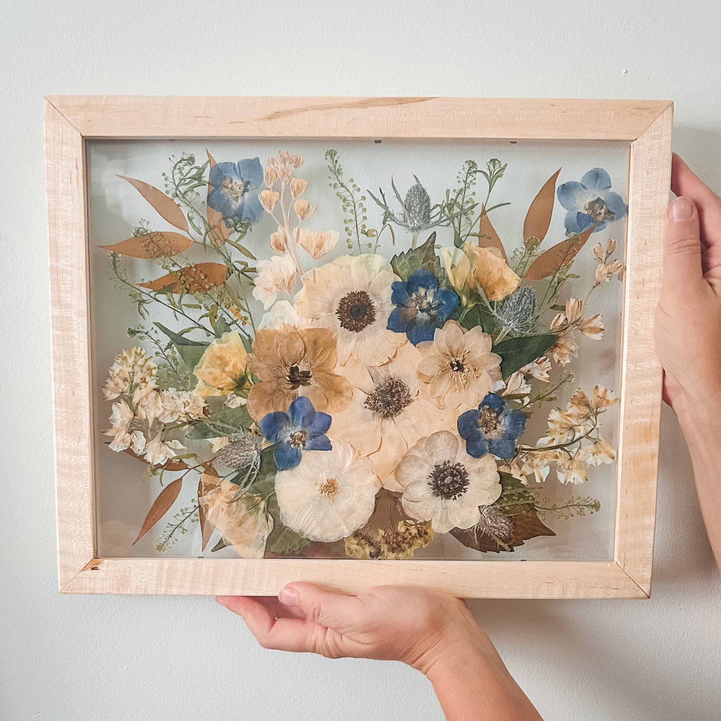 Person holding a framed pressed flower art piece against a white wall.