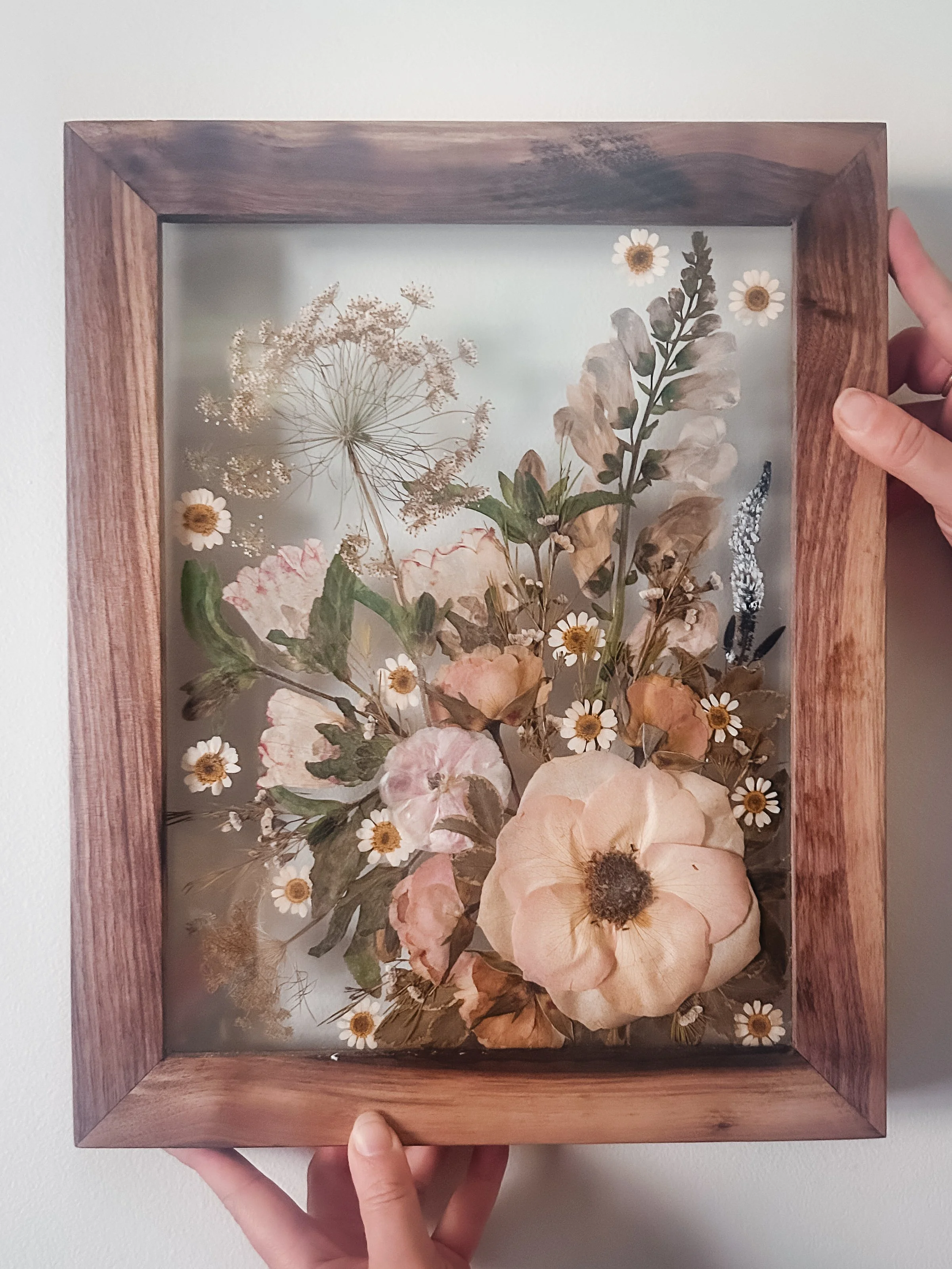 A person holding a wooden framed glass display case with dried flowers and small white daisies arranged inside.