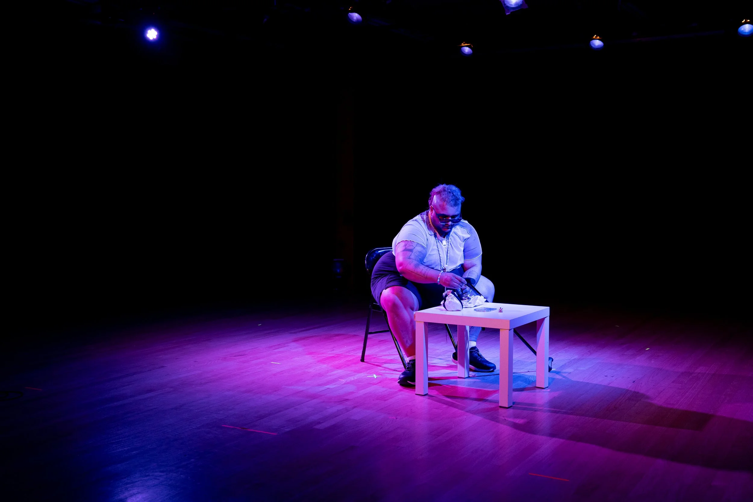 photo of Jai onstage, sitting leaning over a small table. his cane rests against his leg. there is a small bag and a few tiny statues on the table. the lighting is vivid blue and purple, and Jai's skin and hair appears neon purple.