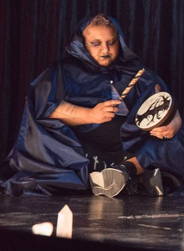 a slightly blurry photo of Jai onstage, sitting with crossed legs. ze is wearing a long navy blue cloak that covers most of his body, and ze holds a drum and a baton.