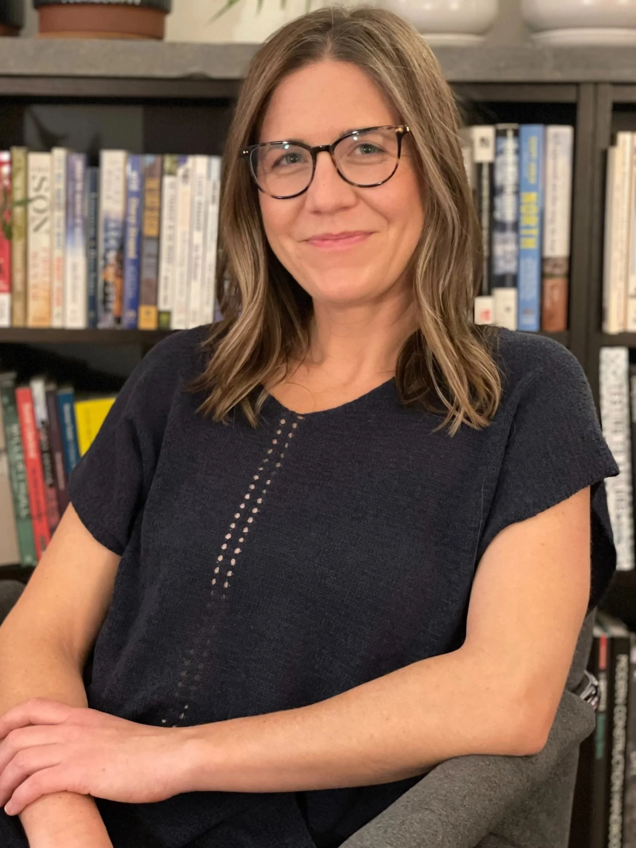 A woman with shoulder-length brown hair, glasses, and a black top sitting in front of a bookshelf.