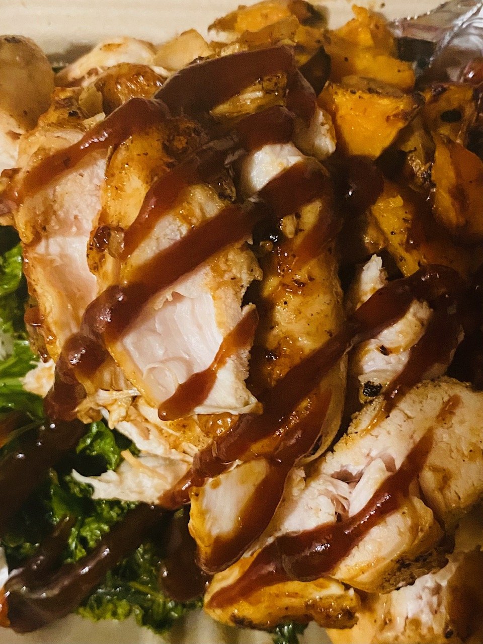 Close-up of sliced grilled chicken breast with dark sauce drizzled on top, served with roasted vegetables and greens.