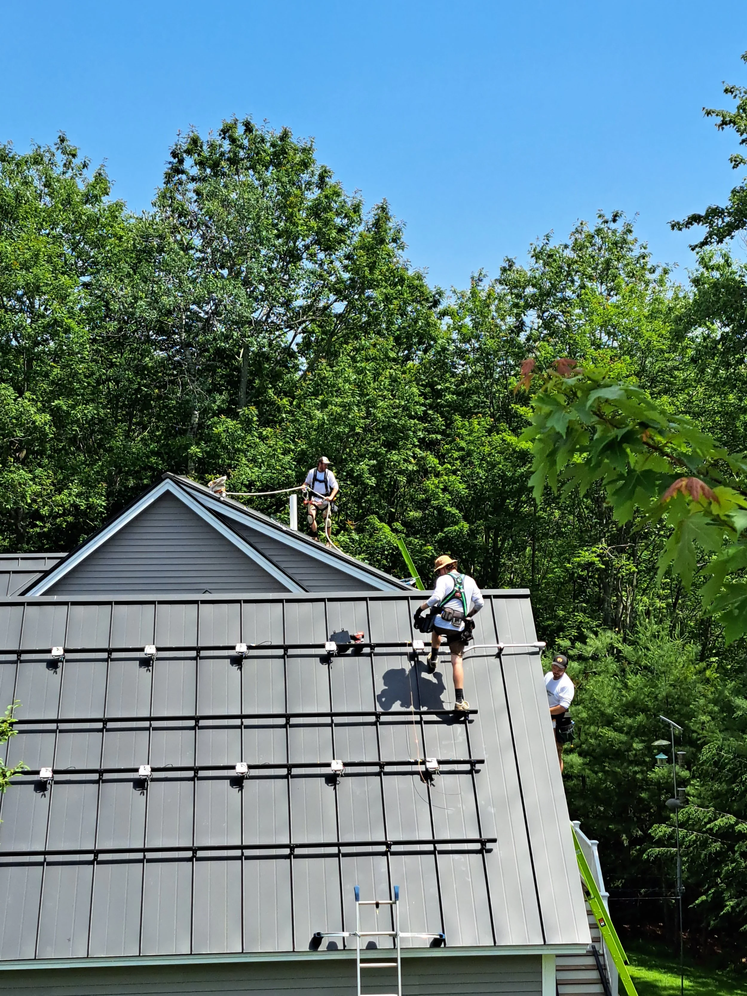 Workers installing solar panel framework on a metal roof with trees in the background.