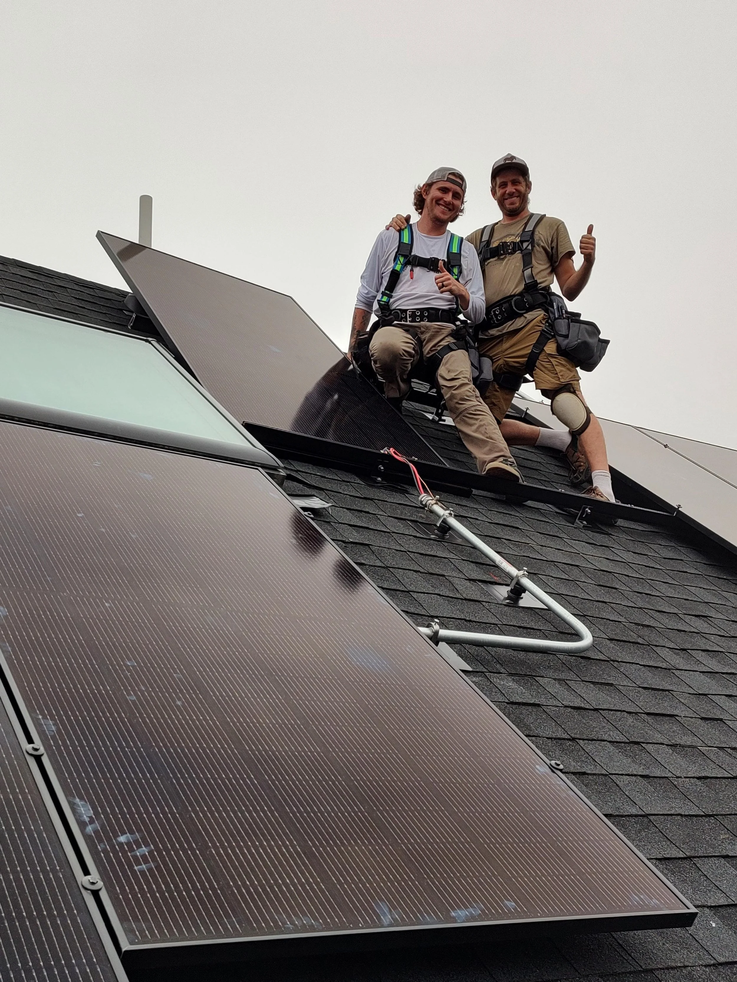 Two workers on a roof with solar panels, wearing safety gear and smiling.