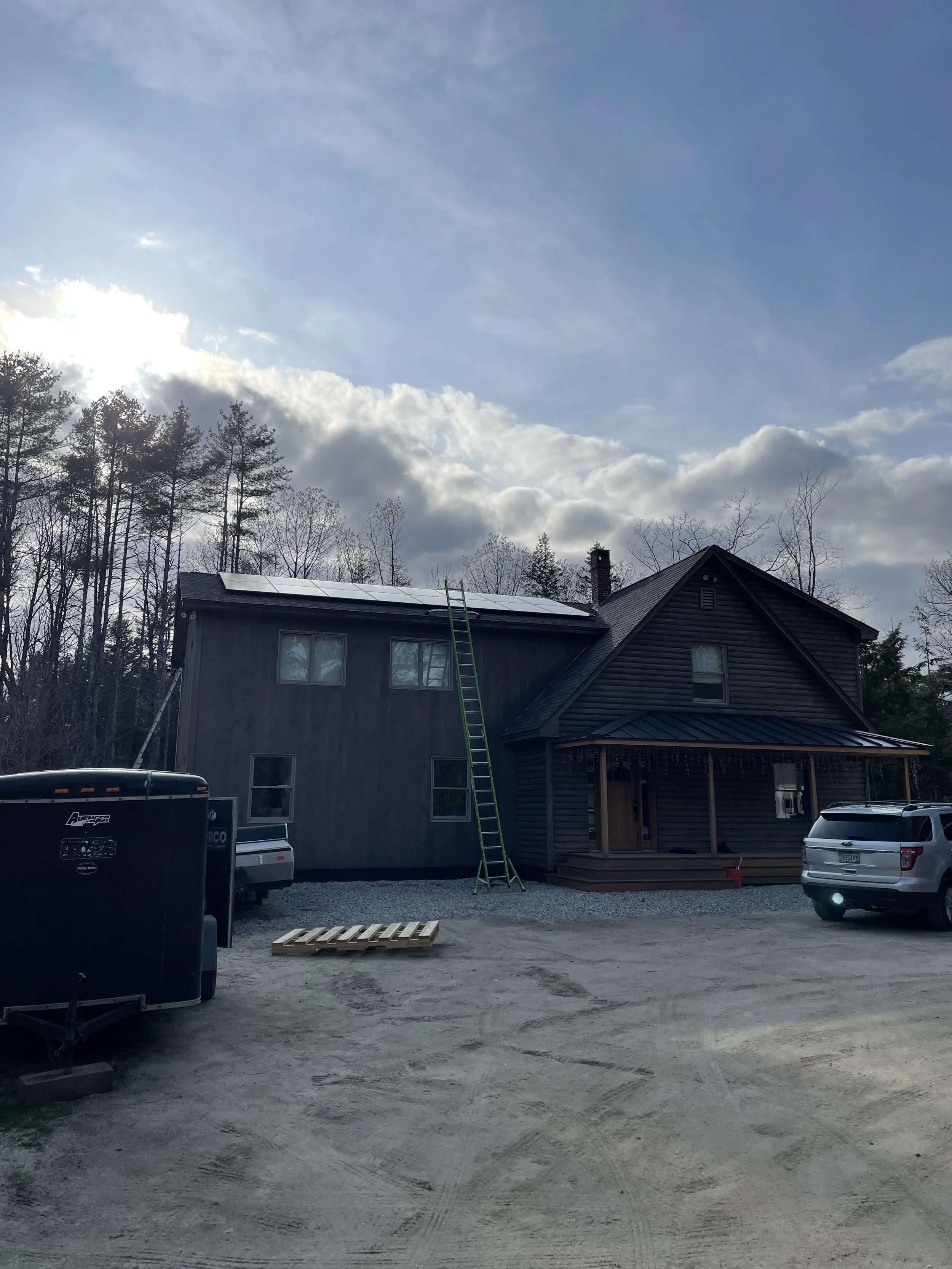 Two-story wooden house with solar panels, a ladder leaning against the roof, surrounded by trees, parked cars, and a small trailer. Overcast sky with sun peeking through clouds.