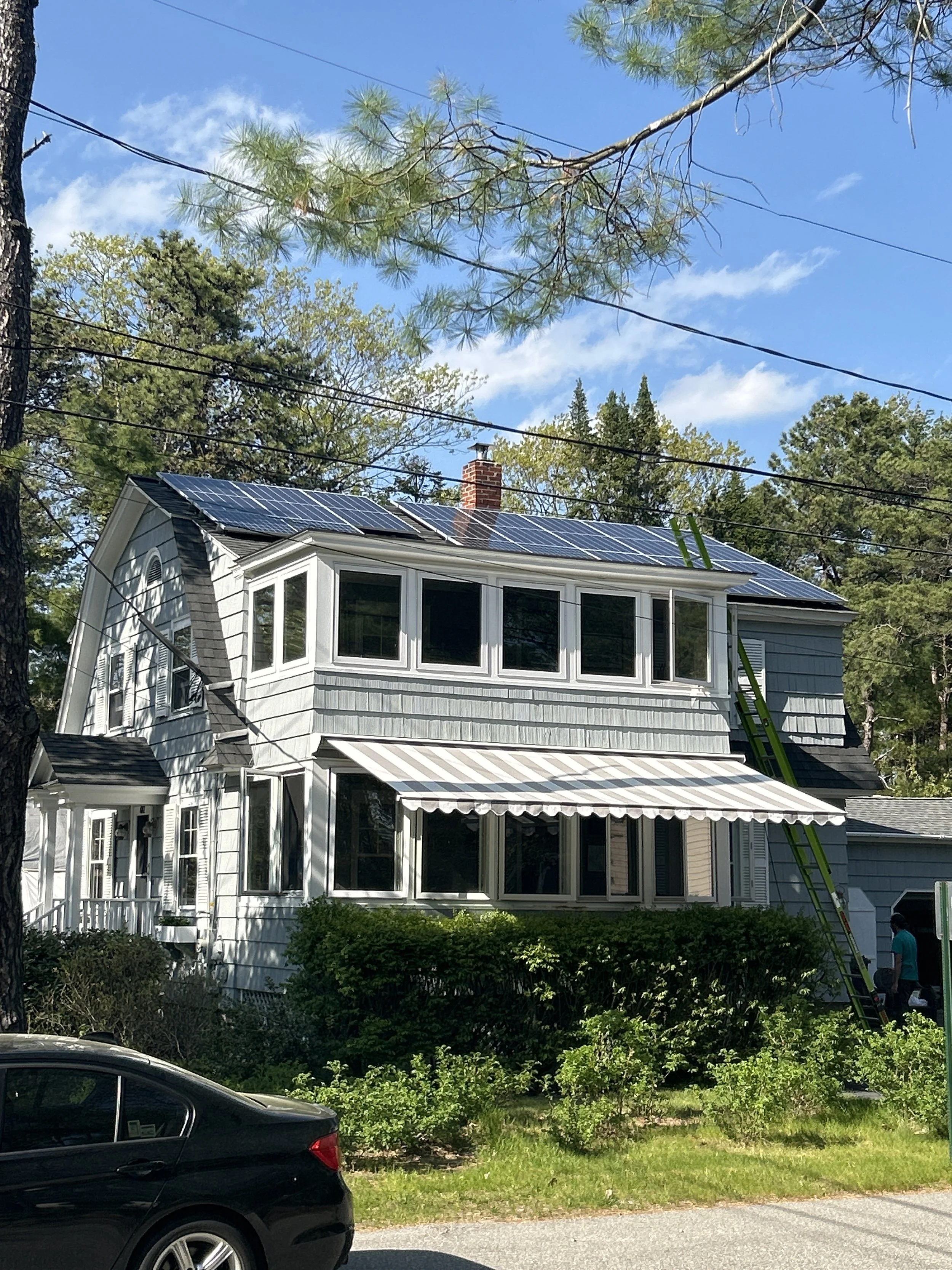 Gray two-story house with solar panels on the roof, surrounded by trees, with a striped awning over the front windows. A ladder is leaning against the house.